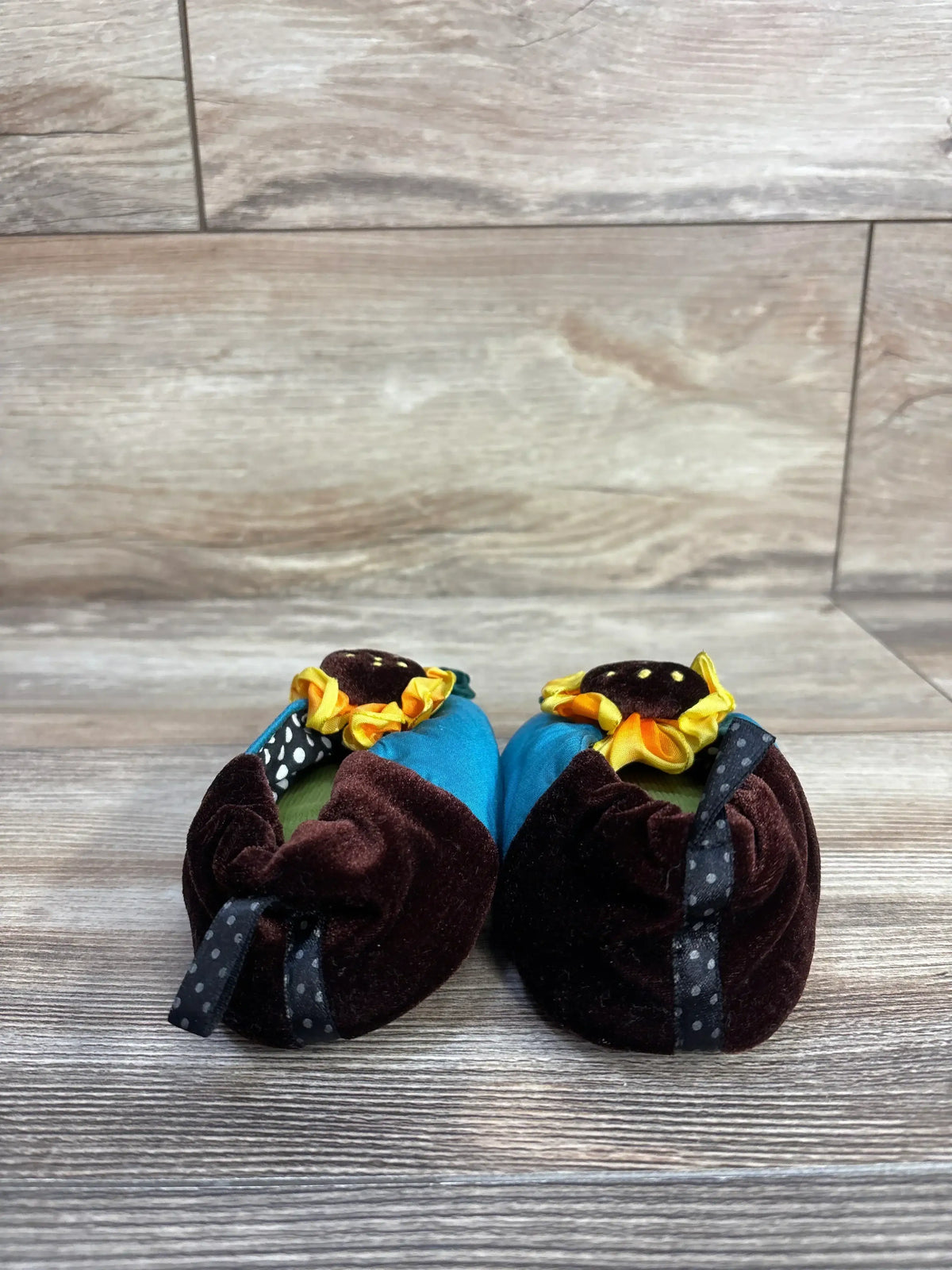 Front view of Goody Sassy Slippers showcasing teal uppers, brown velvet backs, and yellow flower details on a wood-look floor