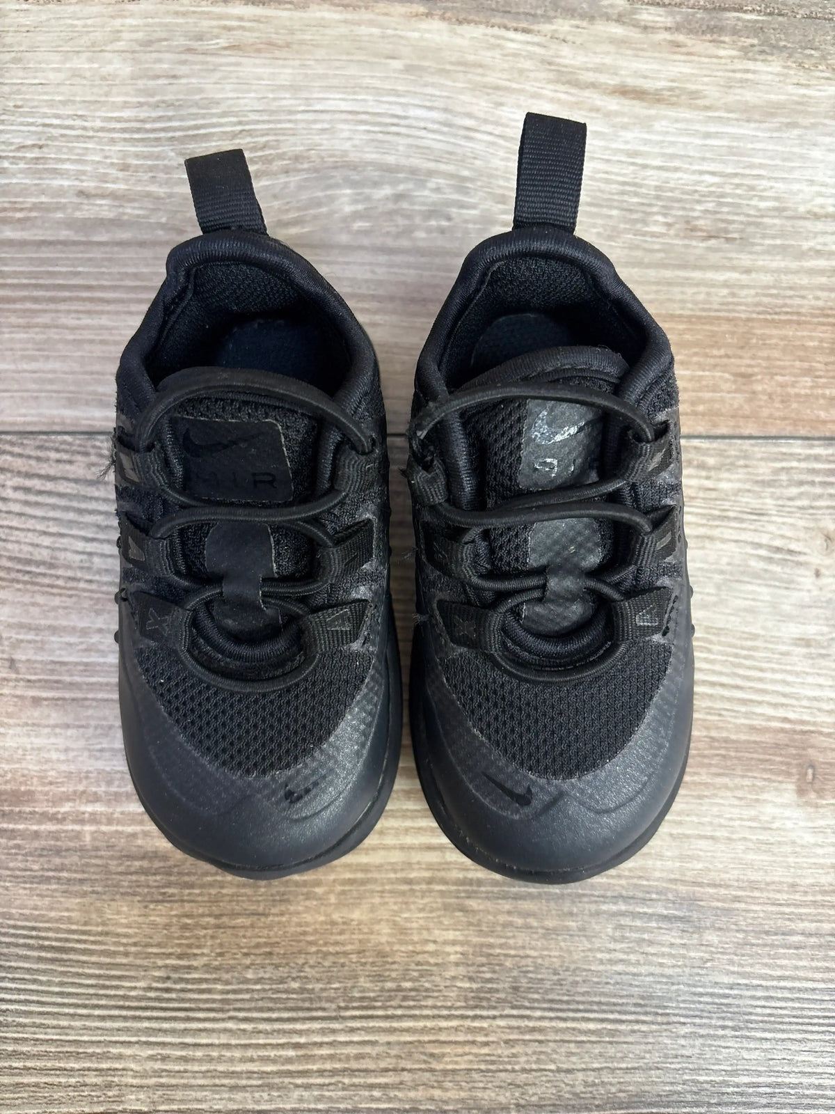Overhead view of Nike Air Max Axis toddler black sneakers showing bungee laces, mesh upper, pull tabs on light wood floor