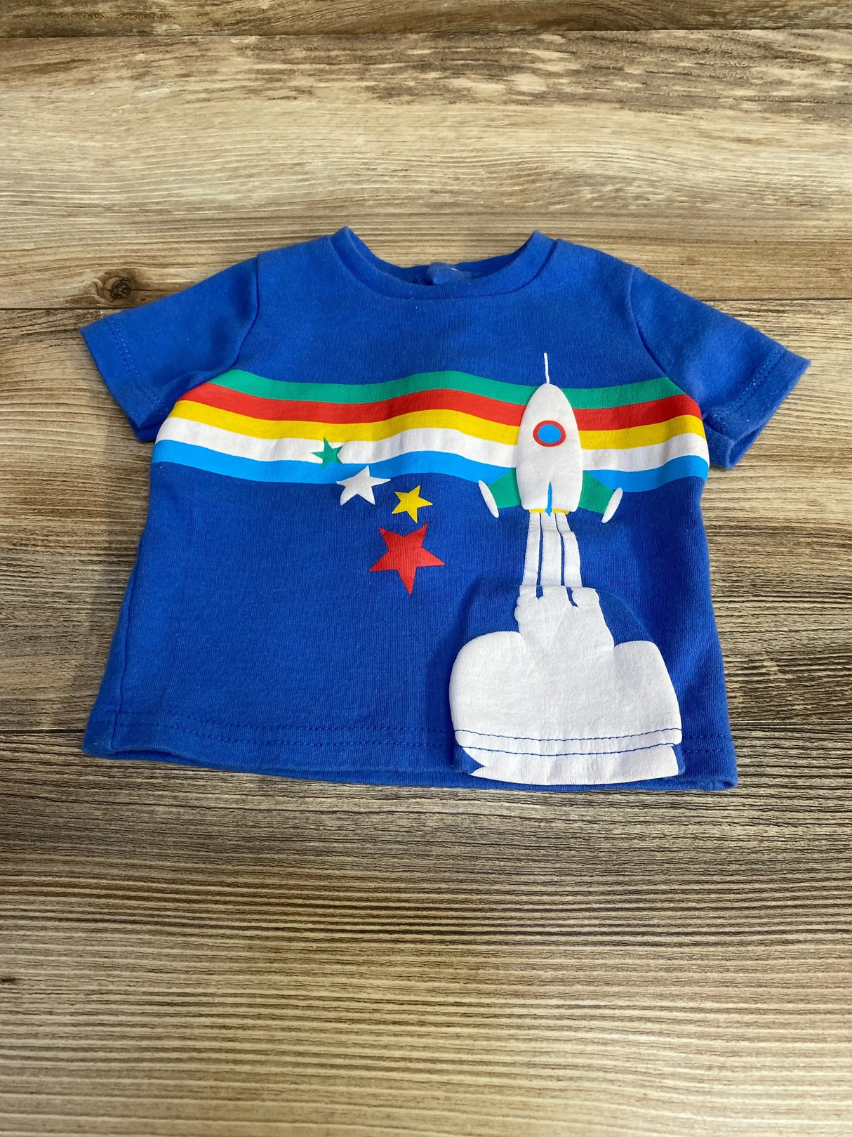 Front view of blue Lily and Jack short-sleeve baby shirt (6-12 months) with white rocket applique and rainbow stripe on wood