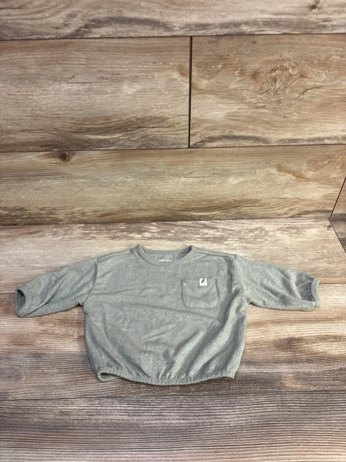 Front view of Easy Peasy long sleeve shirt showcasing soft green color and chest pocket on a wood grain background