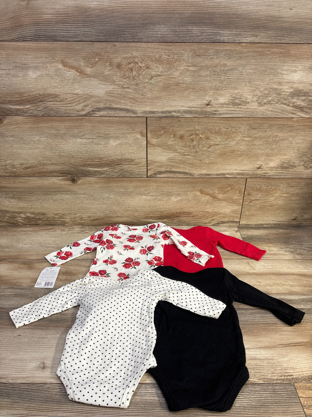 Top-down view of 4 Hudson Baby long-sleeve bodysuits (3-6M) showing solid red, solid black, white with black polka dots, and white with red roses on wood background