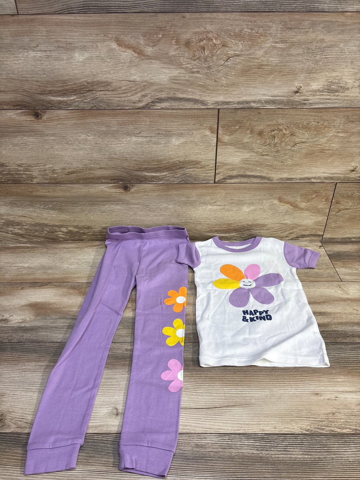 Flat lay of Baby Gap 2-piece pajamas (5T) showing white tee with smiling multicolor daisy and HAPPY & KIND text and purple leggings with flower prints on wood background