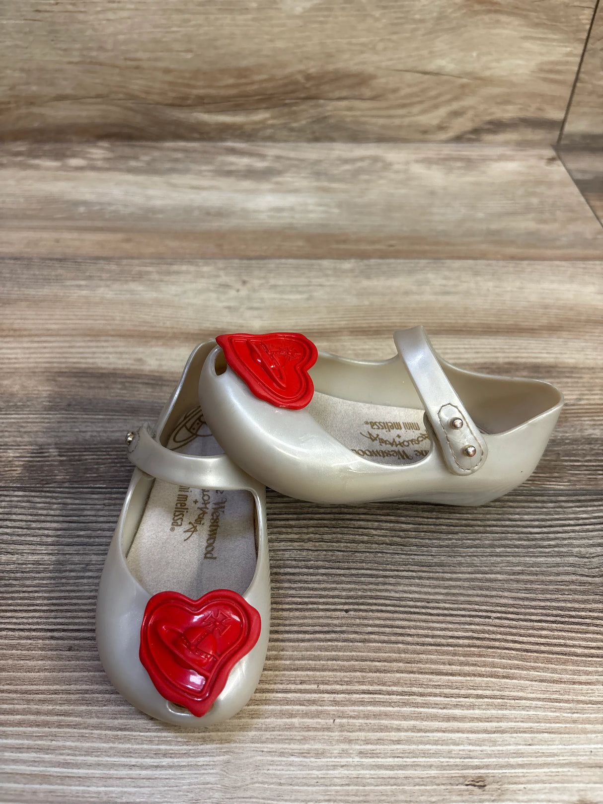 Angled top view of Vivienne Westwood x Mini Melissa Mary Jane shoes in pearlized cream with red heart charms on wood background