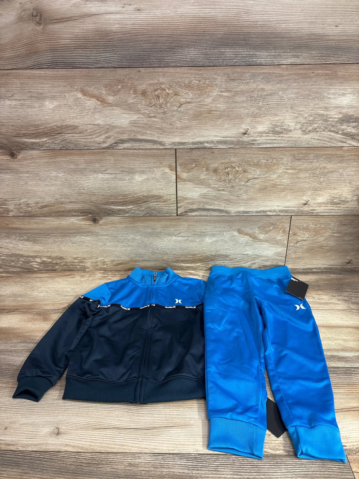 Flat lay Hurley 2-piece tracksuit size 4T: zip jacket blue/black and bright blue pants with elastic cuffs on wood floor