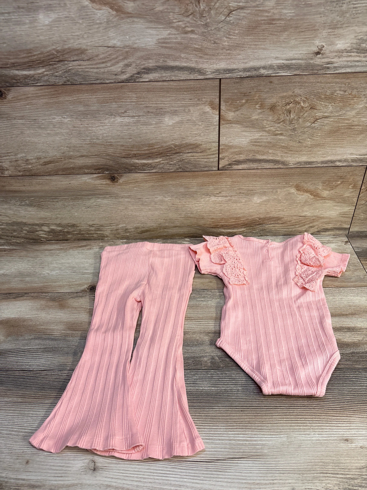 Flat lay of pink ribbed baby bodysuit and matching flare pants showcasing eyelet lace ruffle sleeves on wood background