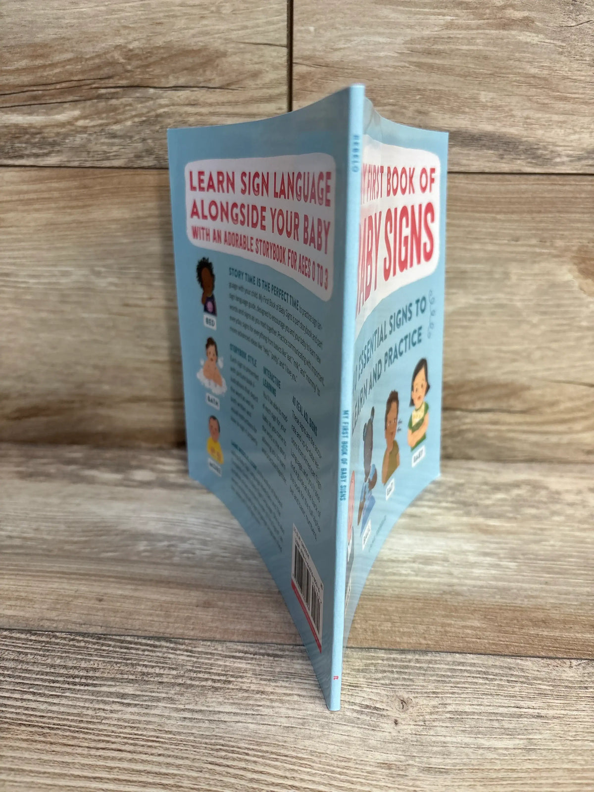 Three quarter view of First Book of Baby Signs paperback standing upright showcasing blue cover and back illustrations on wood plank floor