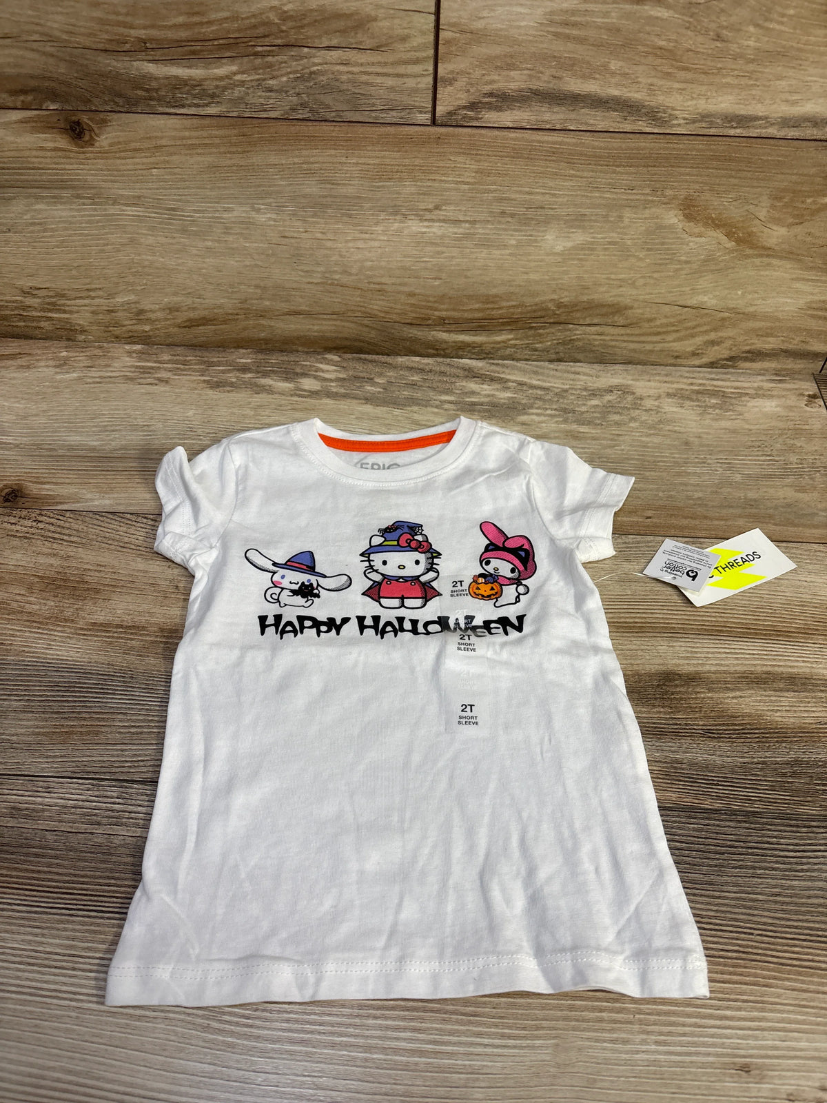 Front flat lay of Epic Threads x Hello Kitty white toddler T-shirt 2T with Halloween character graphic and Happy Halloween text on wood floor