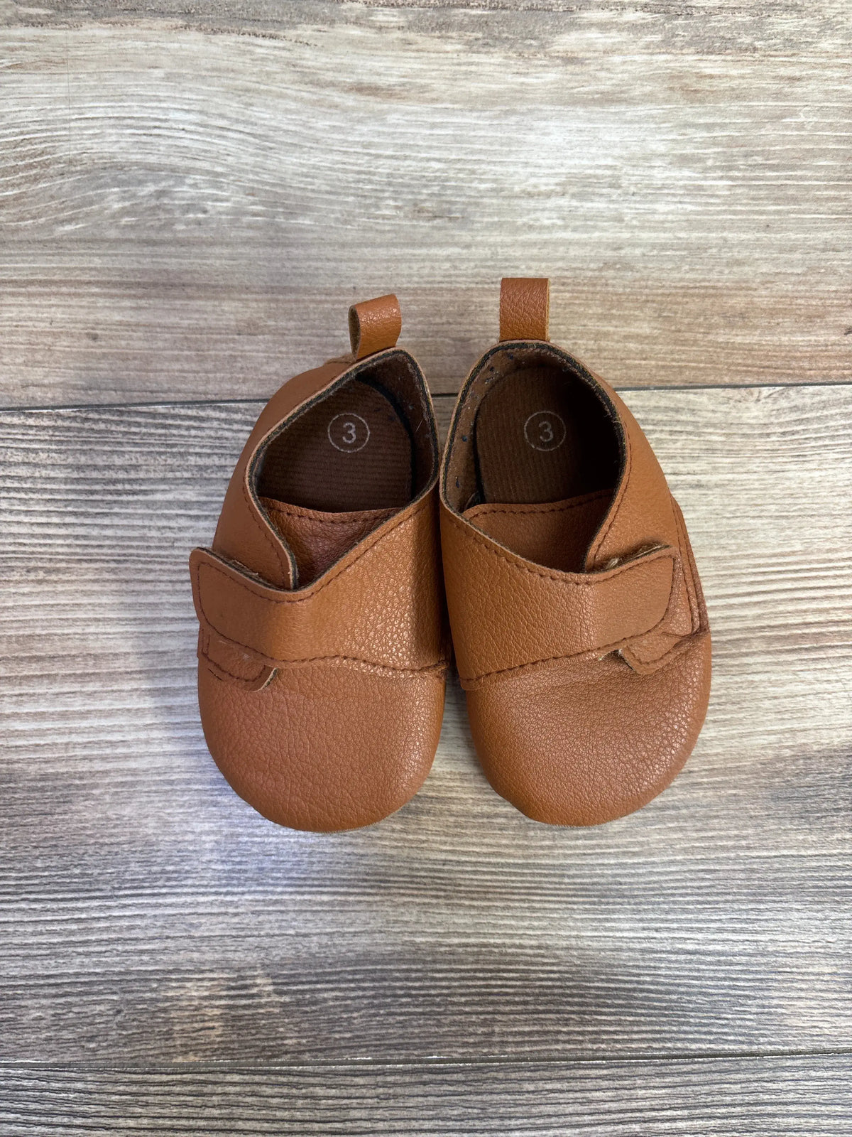 Top view of brown faux leather first walking soft sole shoes showing hook and loop closures with pull tabs on wood grain