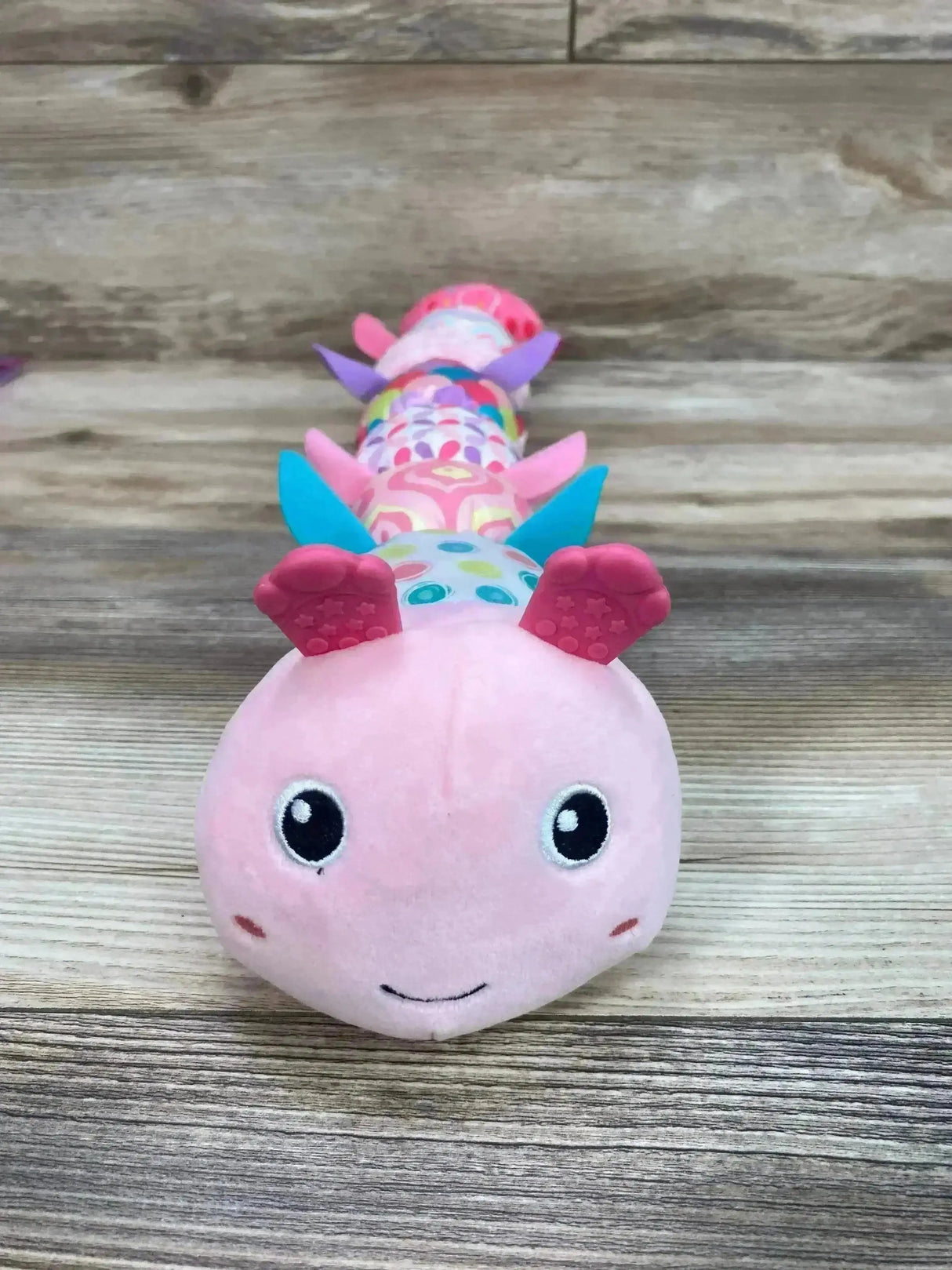 Front view of pink Sumobaby musical caterpillar plush with textured silicone feet and colorful patterned segments on wood background