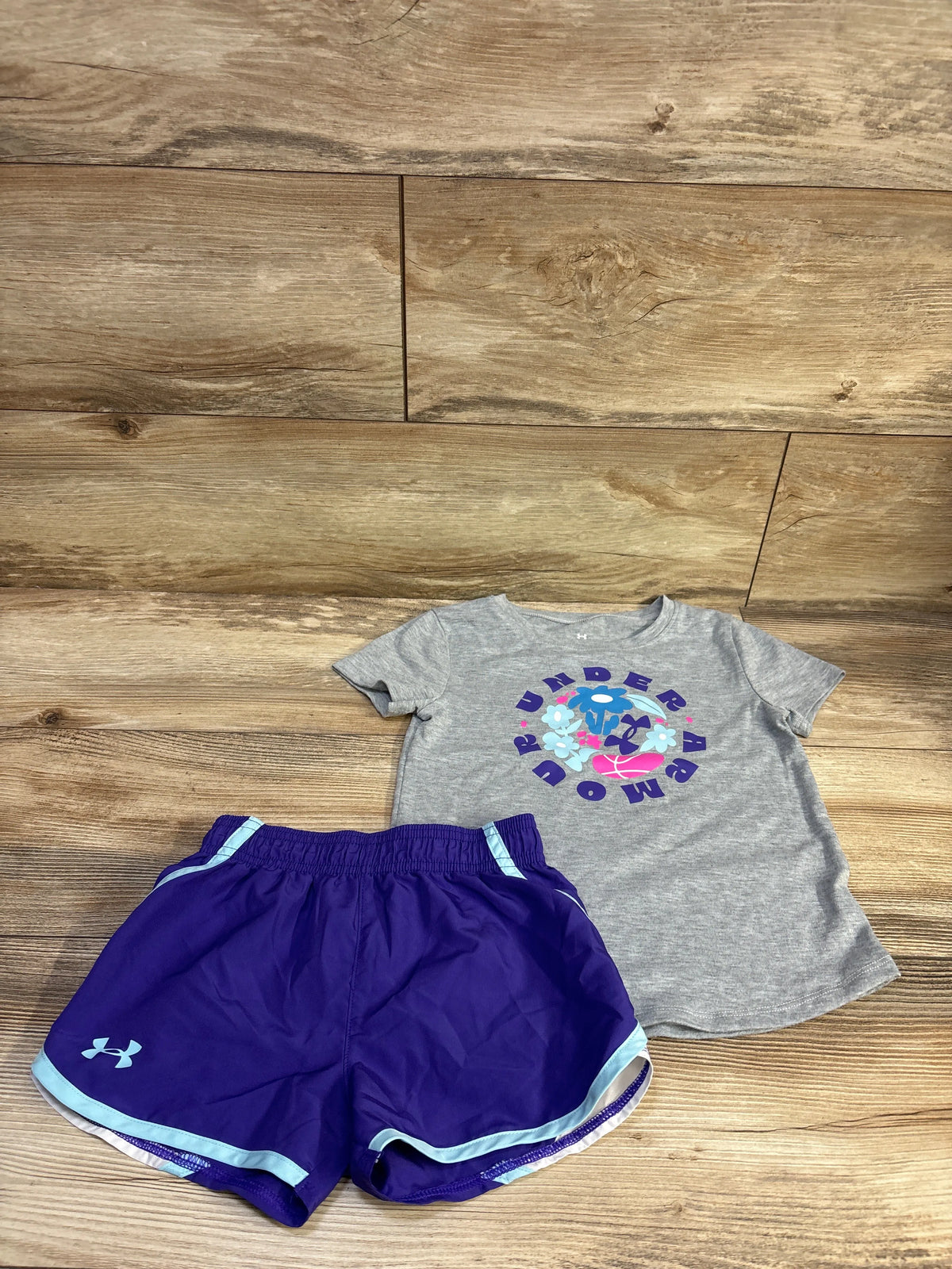 Top-down view of Under Armour 2pc shirt and shorts set showcasing a gray graphic shirt, purple shorts, and blue trim on wood