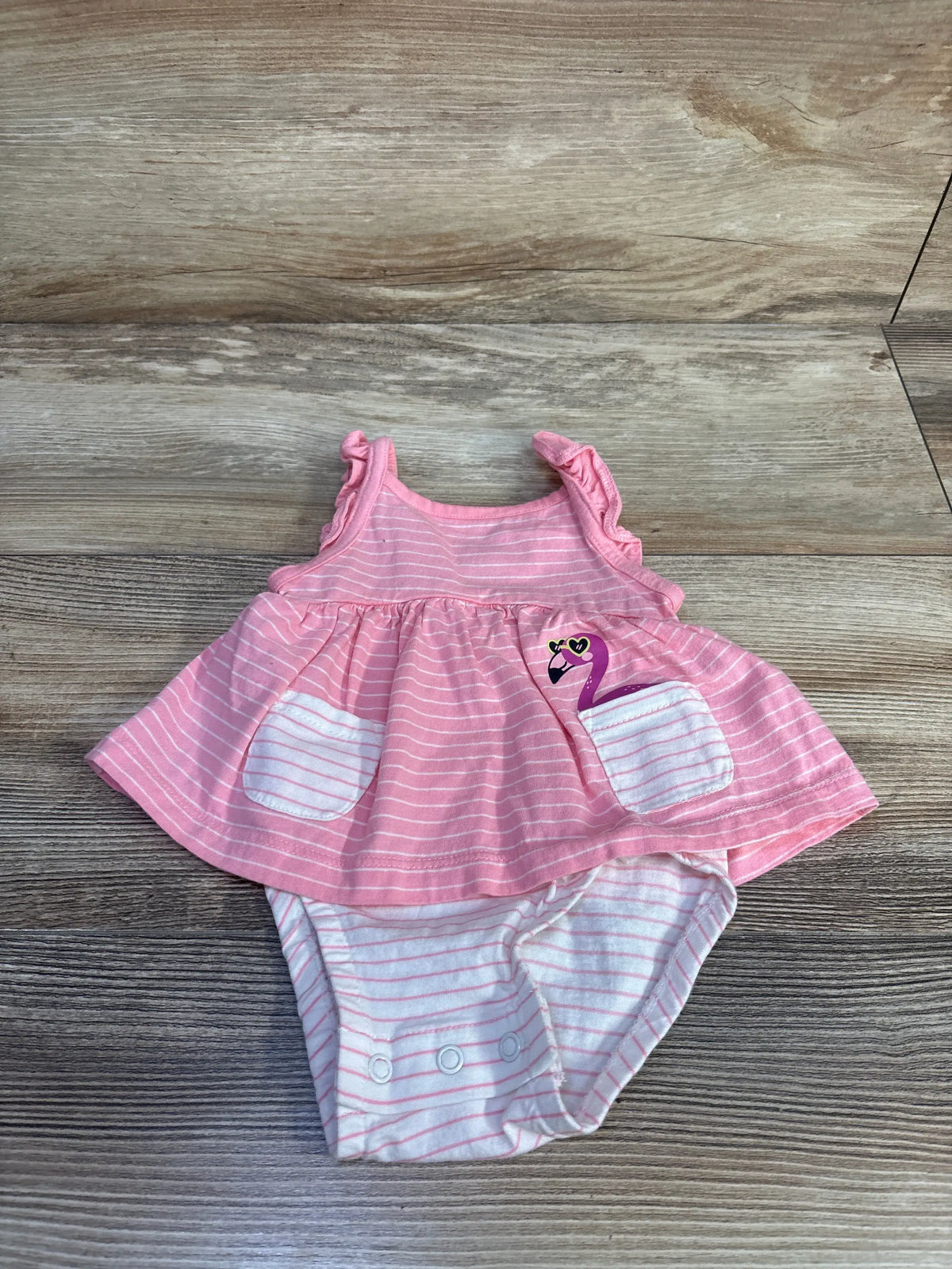 Front view of Carter’s pink flamingo bodysuit dress showcasing its sleeveless design, striped skirt and snap closures on a wood surface
