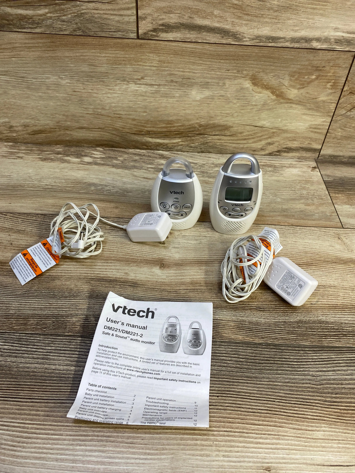 Front view of two white VTech DM221 baby monitor units with LCD and buttons, two power adapters and manual on wood background