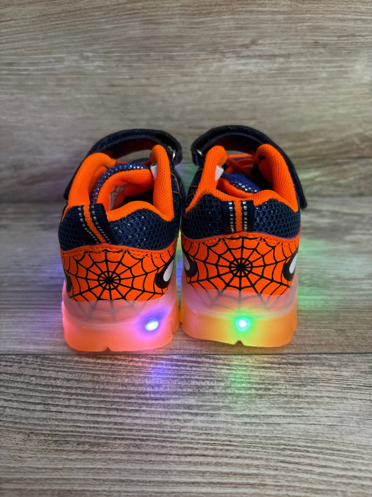 Rear view of navy and orange toddler light-up sneakers showing spiderweb heel print and multicolor LED soles on wood floor