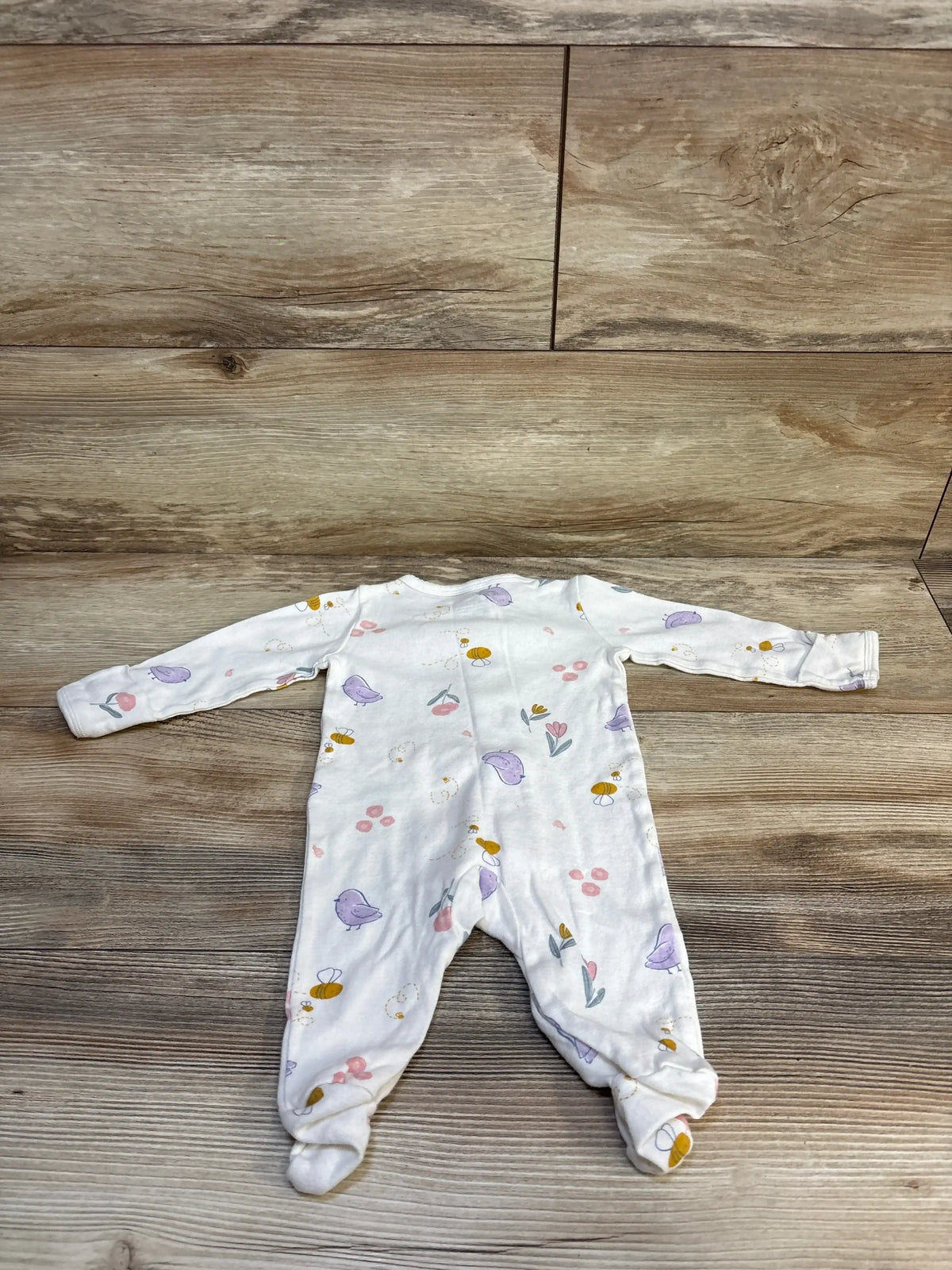 White baby onesie with purple birds, flowers, and bees on wooden floor