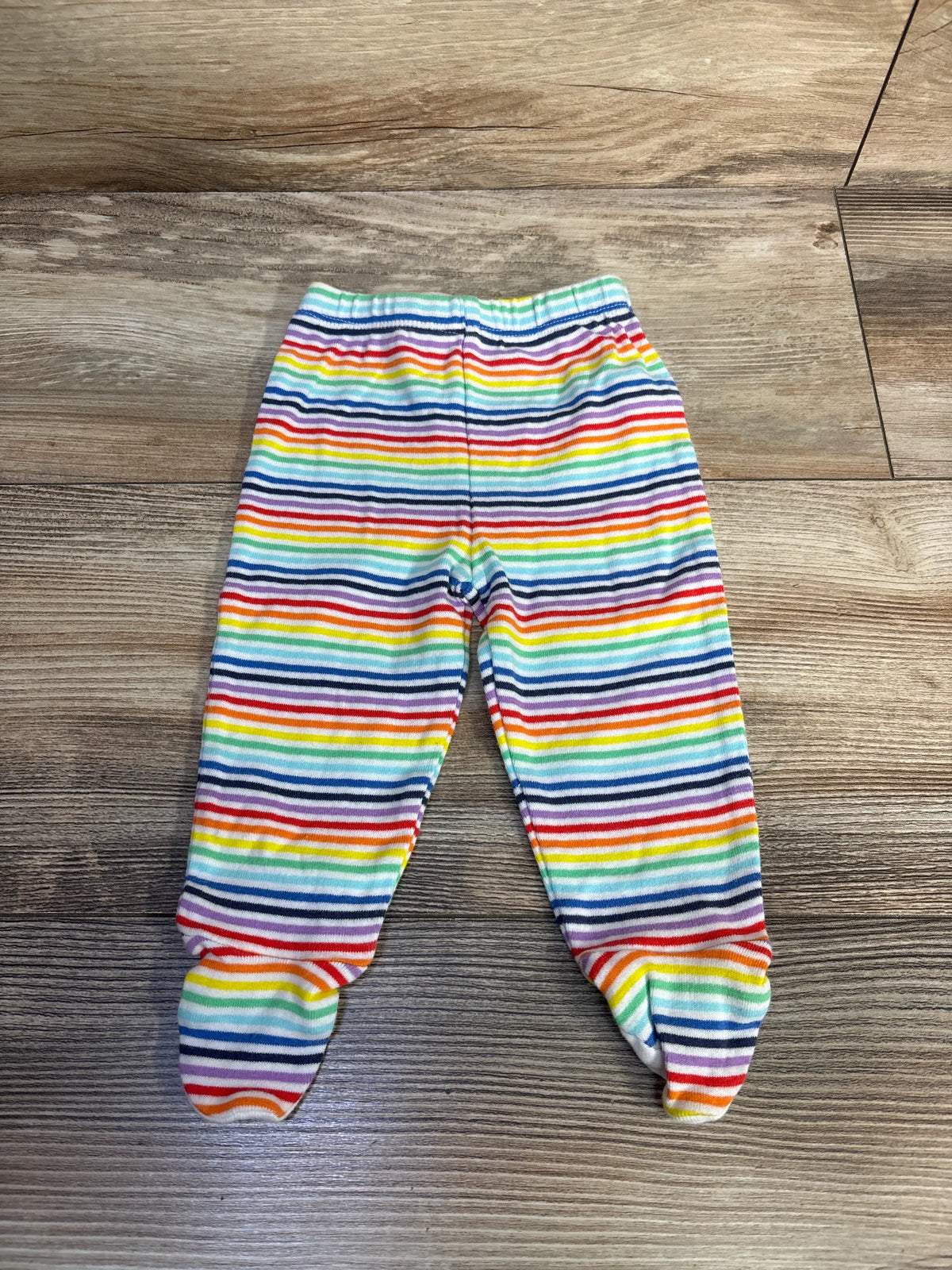 Top-down view of Primary striped footed pants, size 0-3 months, showcasing rainbow stripes and footed design on a wood floor