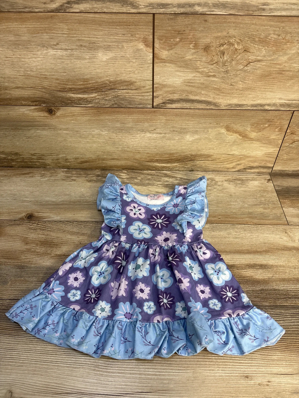 Front view of Pete + Lucy blue floral dress showcasing ruffled sleeves and hem on a wood plank background
