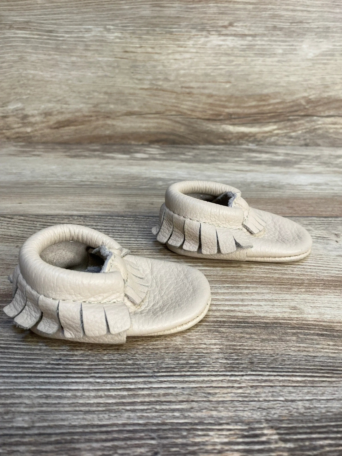 Beige leather baby moccasins with fringe on wooden background, toddler shoes