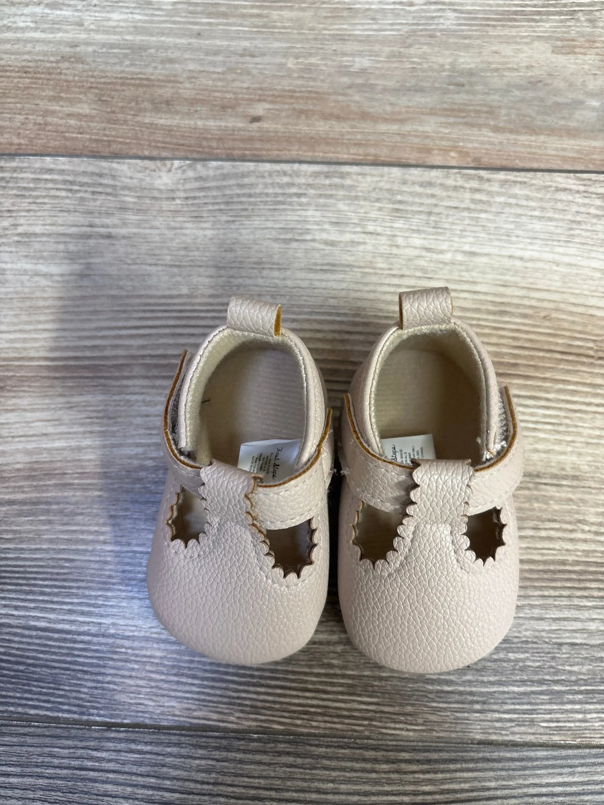 Top-down view of beige First Steps soft-sole baby shoes showing scalloped T-strap and star-shaped cutouts on gray wood floor