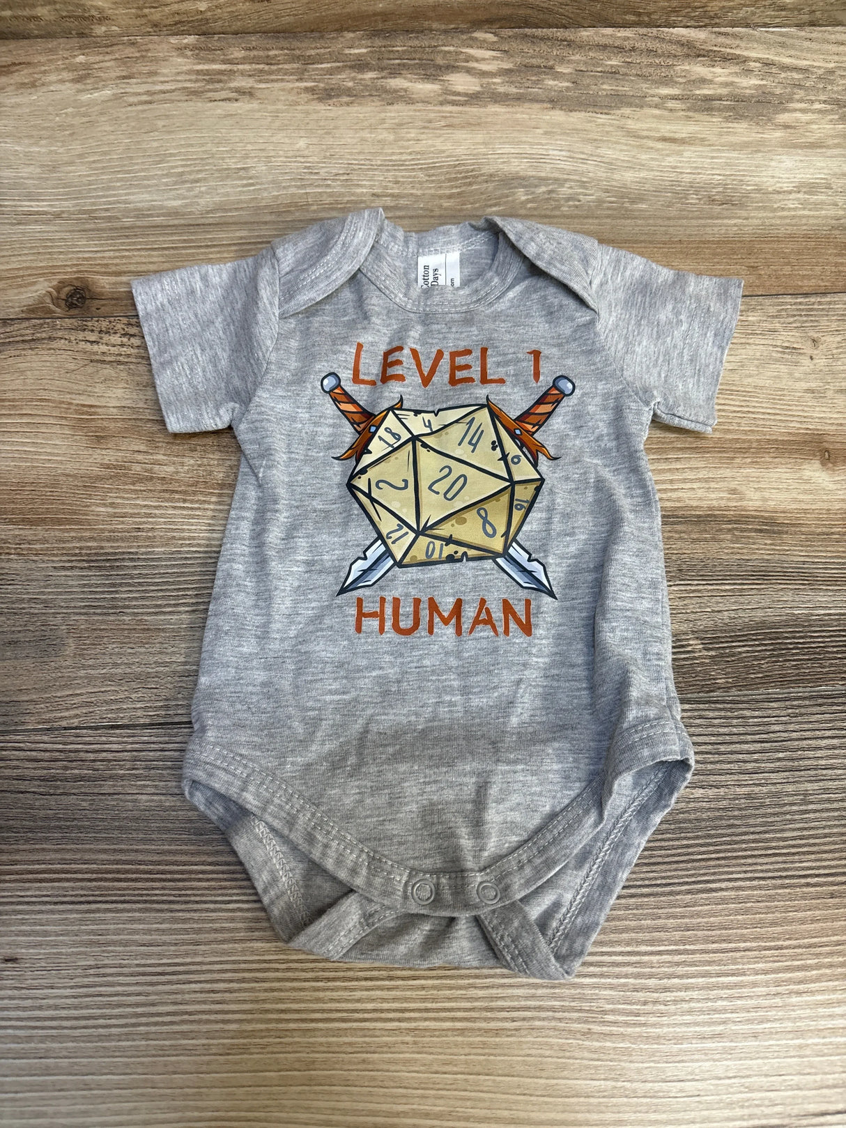 Gray baby onesie with D20 dice, swords, and 'Level 1 Human' graphic, laid on wood background