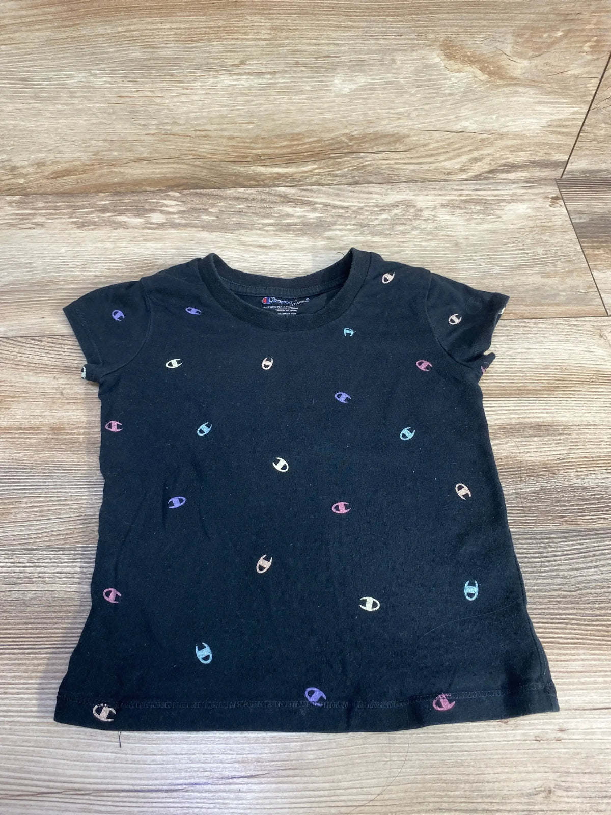 Flat lay of black Champion toddler T-shirt 4T showing scattered small multicolor Champion C logos on wood floor