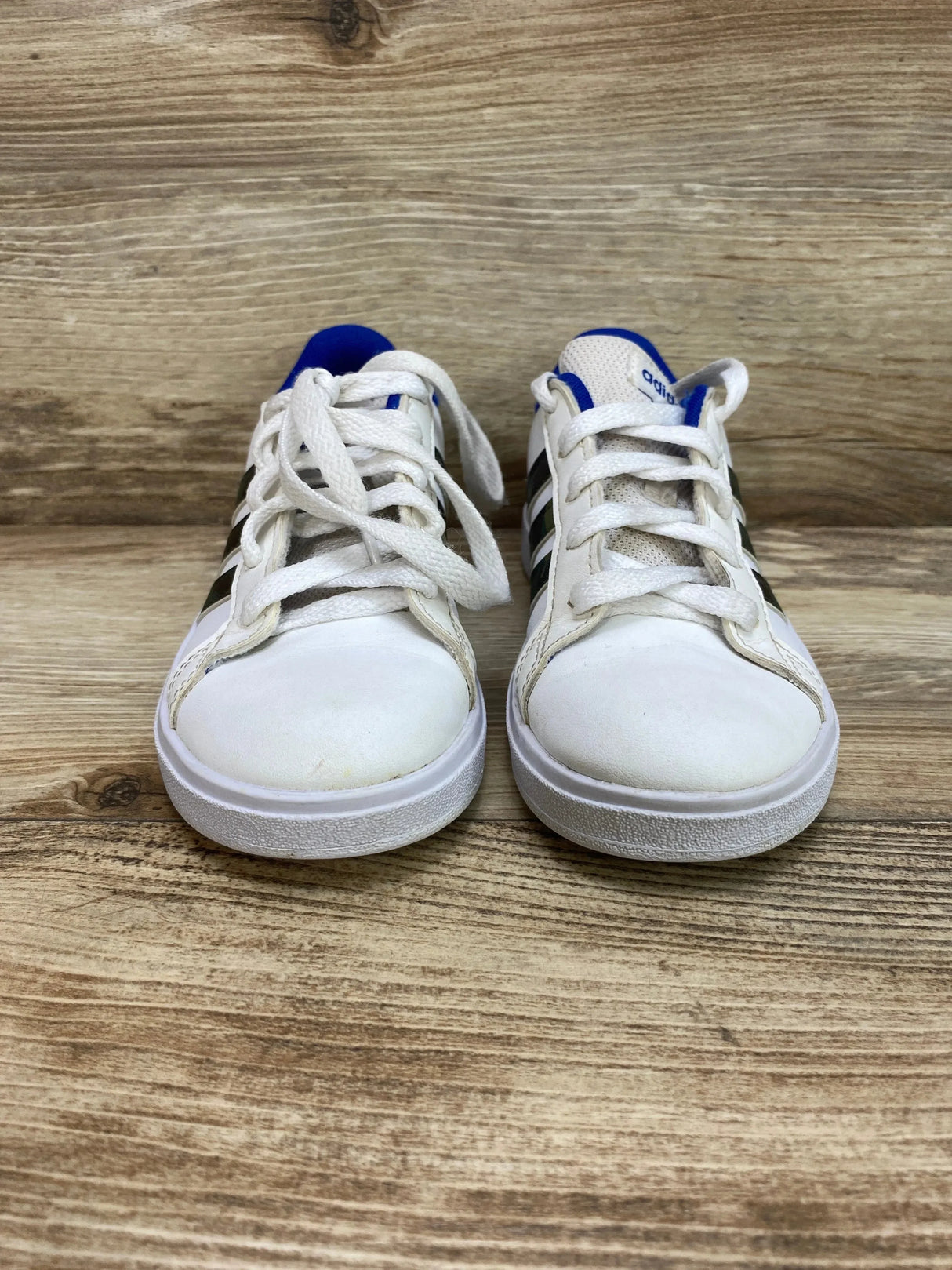 Front view of Adidas toddler sneakers size 12C, white with black three stripes and royal blue heel lining on wood background