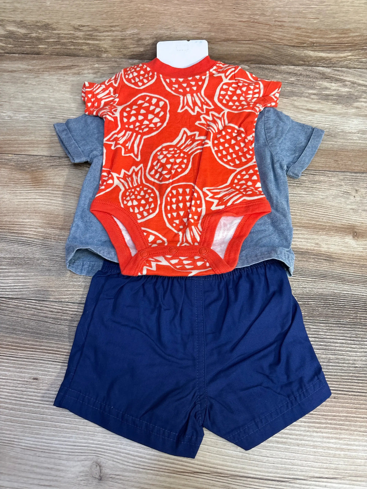 Top-down of 3-piece baby outfit: orange pineapple-print onesie over gray chambray shirt with navy shorts on wooden floor
