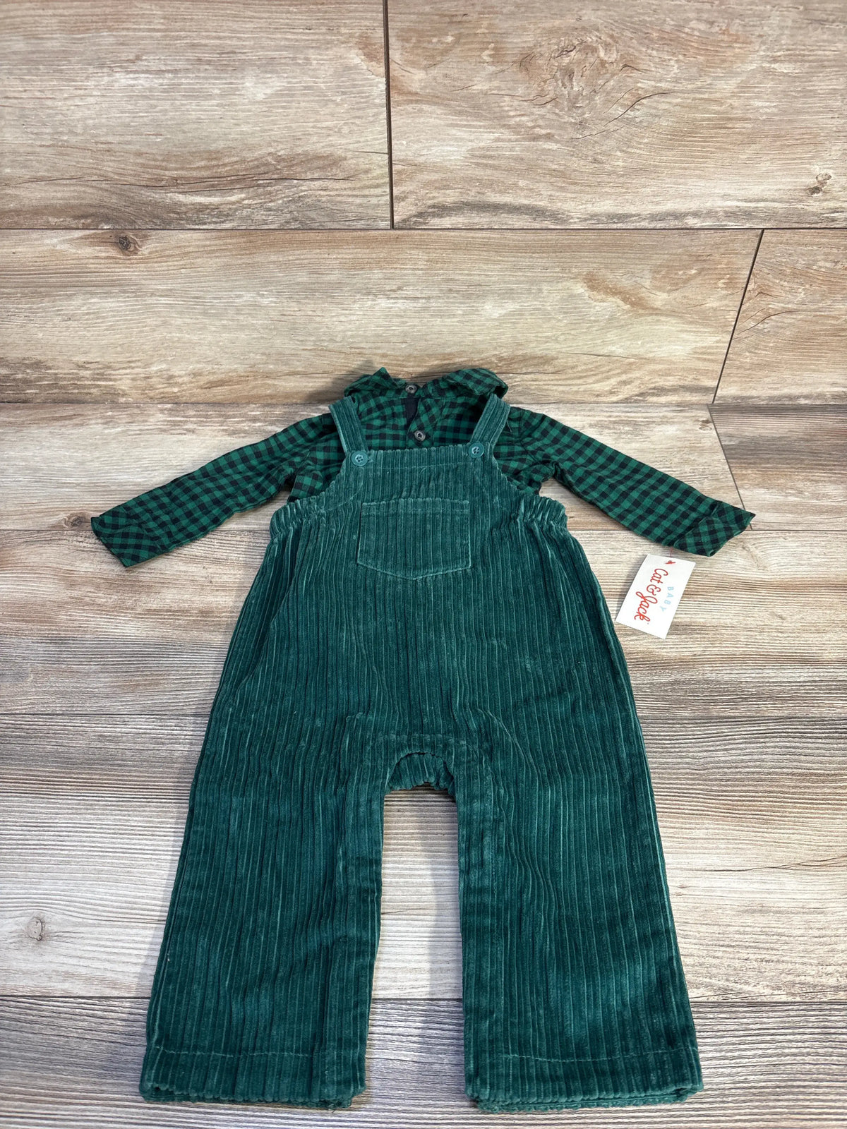 Front flat lay of green corduroy overalls with chest pocket and adjustable straps paired with green plaid button up shirt on wood floor