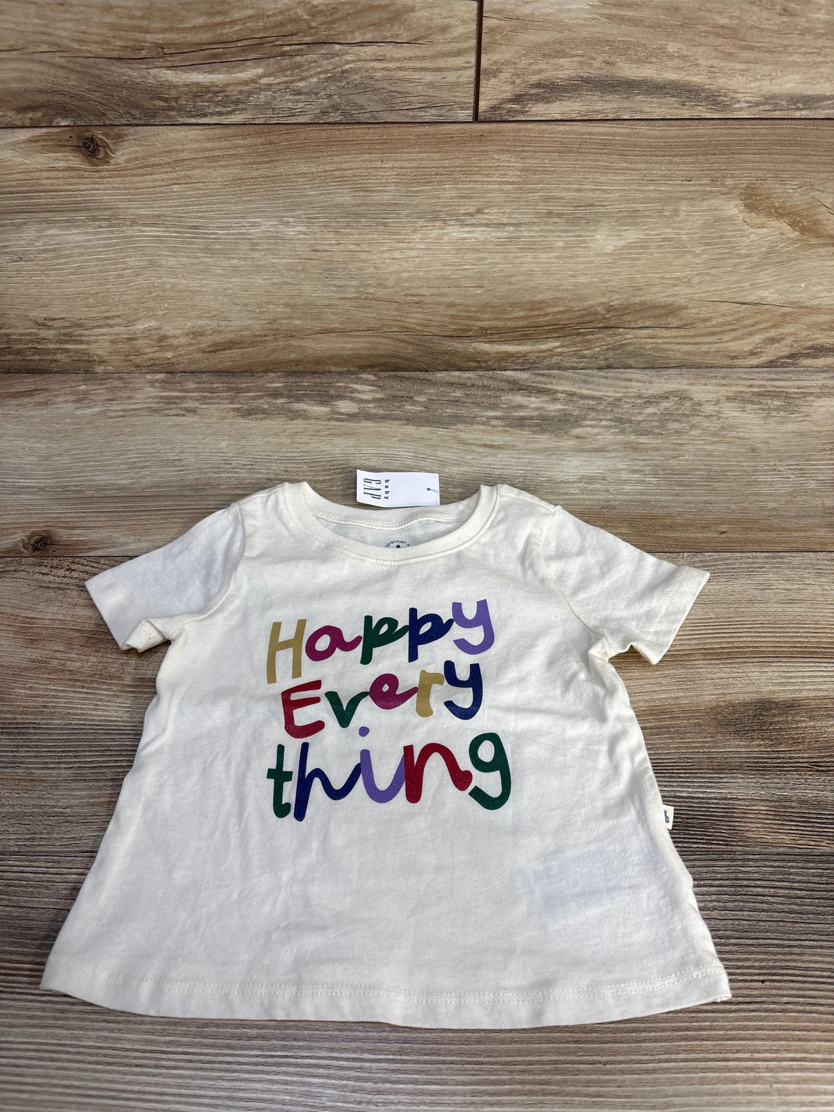 Front view of Baby Gap ivory short-sleeve tee 6-12M showcasing multicolored Happy Everything text on wood background