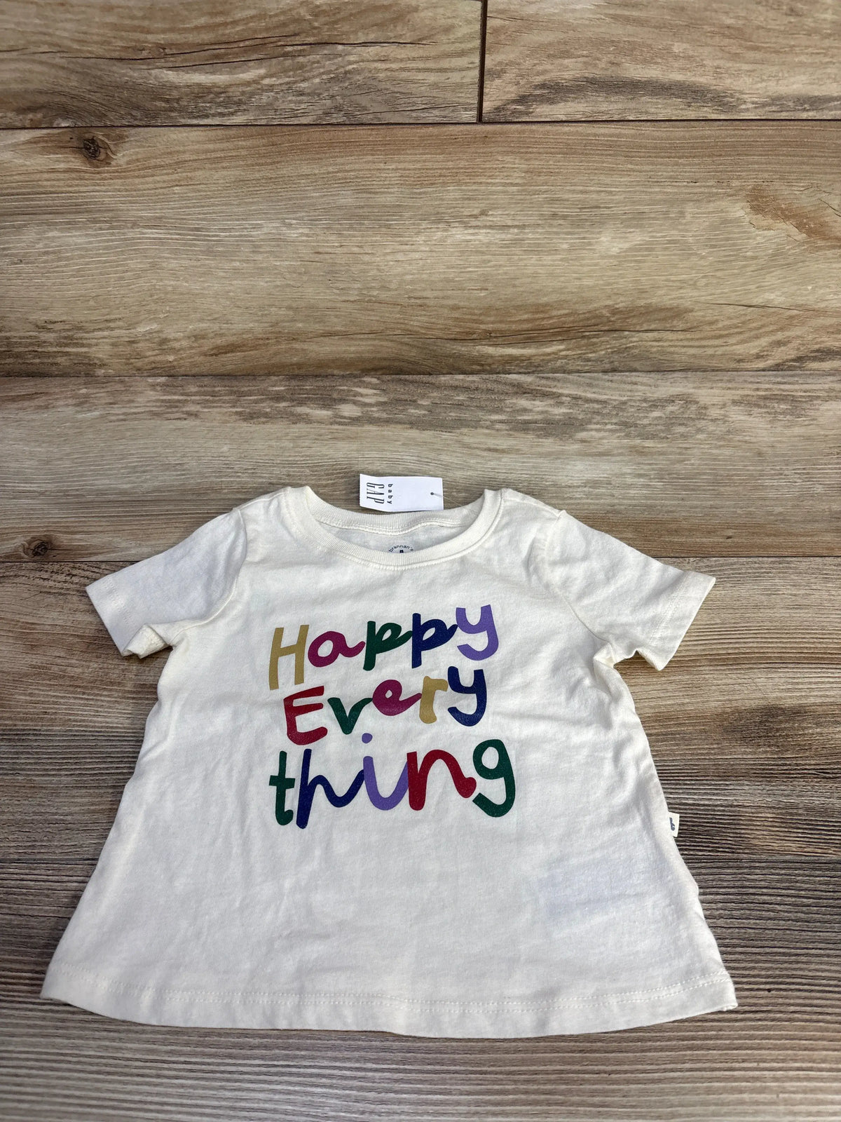 Front view of Baby Gap ivory short-sleeve tee 6-12M showcasing multicolored Happy Everything text on wood background