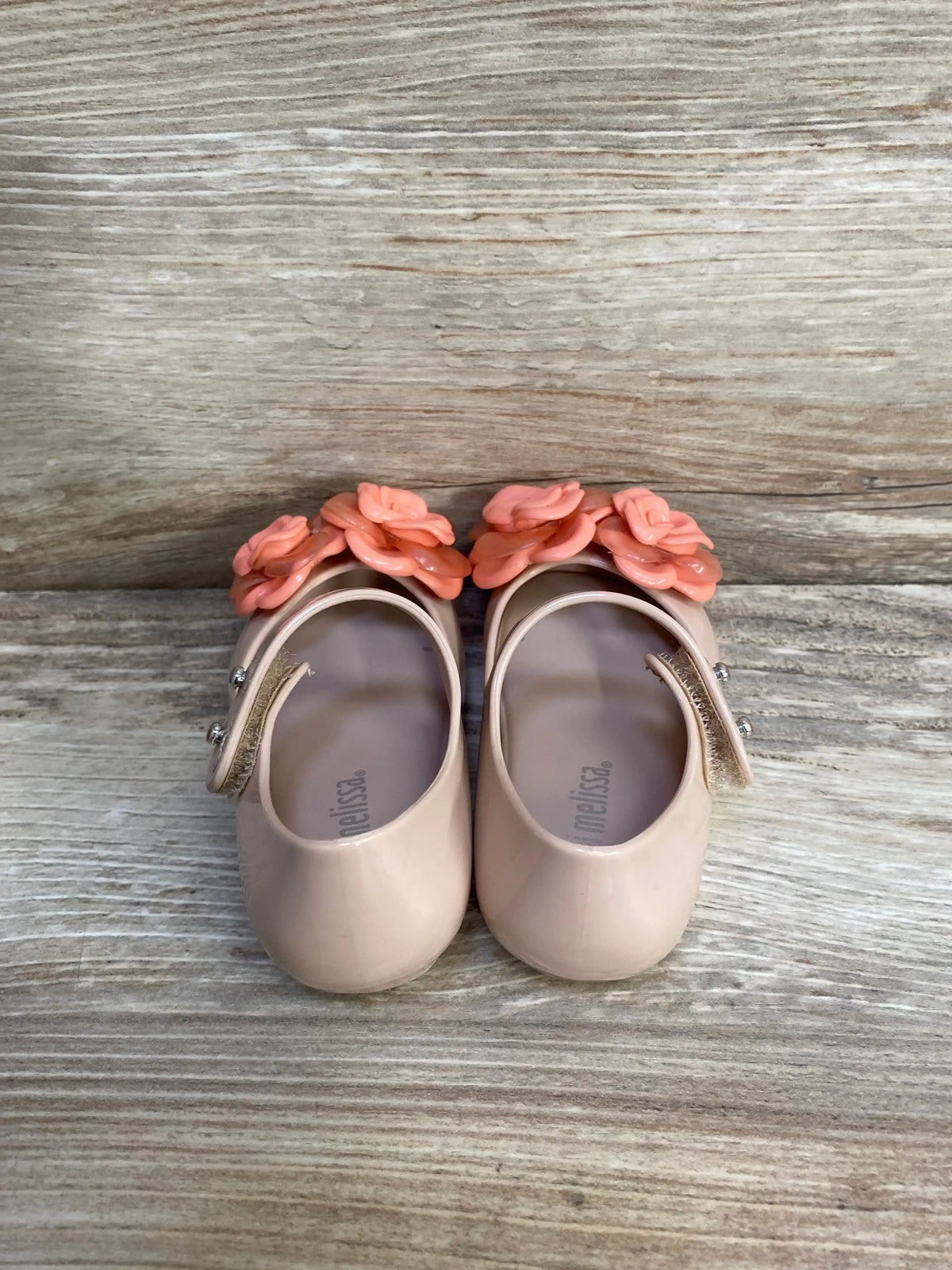 Top view of Mini Melissa Ultragirl flats in beige with coral-pink flower decorations and Mary Jane strap on light wood background
