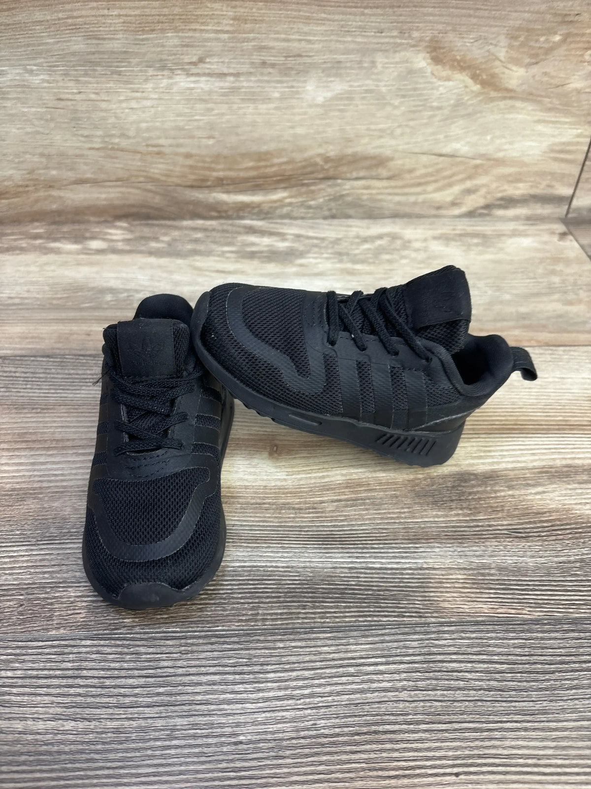 Three-quarter view of Adidas Multix I Triple Black sneakers size 6c showcasing mesh upper and three-stripe side panels on wood plank floor