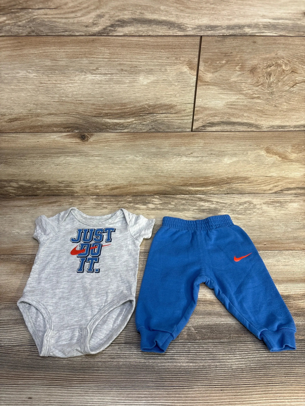 Nike 2-piece baby set flat-lay: grey bodysuit with ‘JUST DO IT.’ text and red swoosh, blue pants with red Nike logo on wood floor