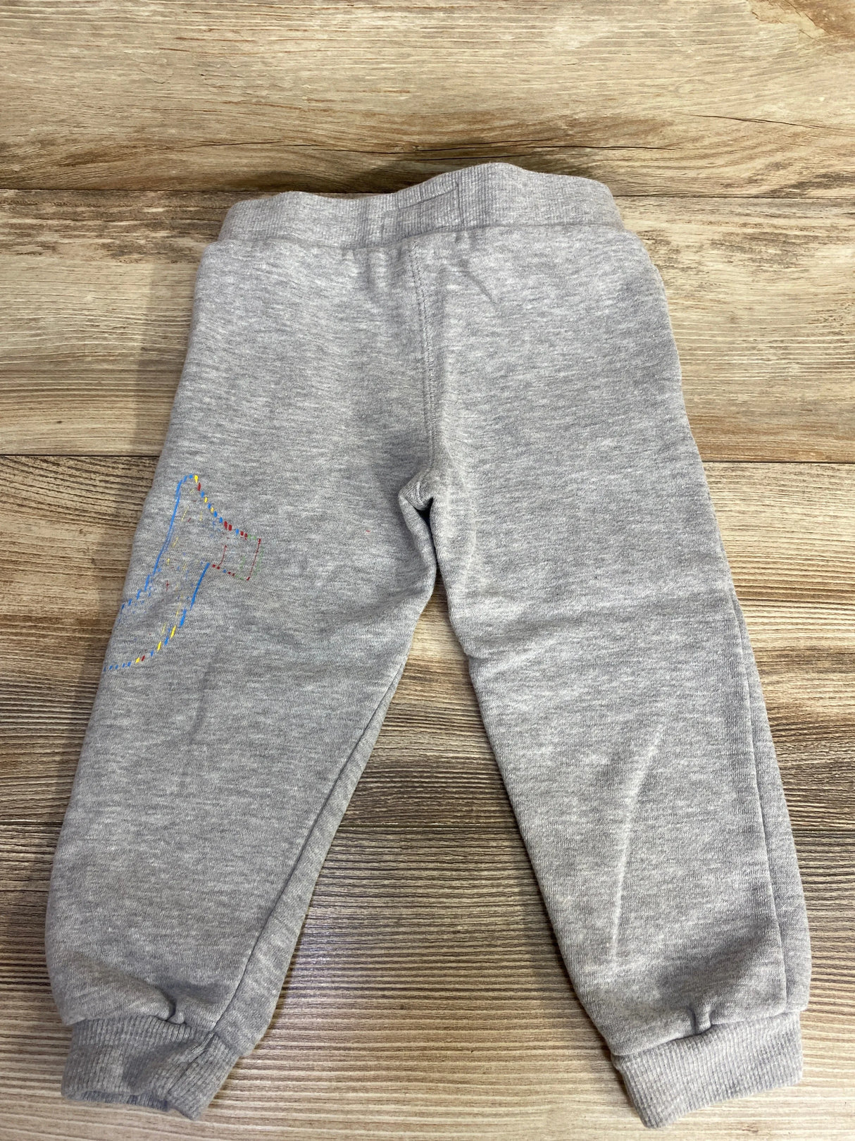 Gray toddler sweatpants with elastic cuffs and colorful graphic on left leg on wood floor