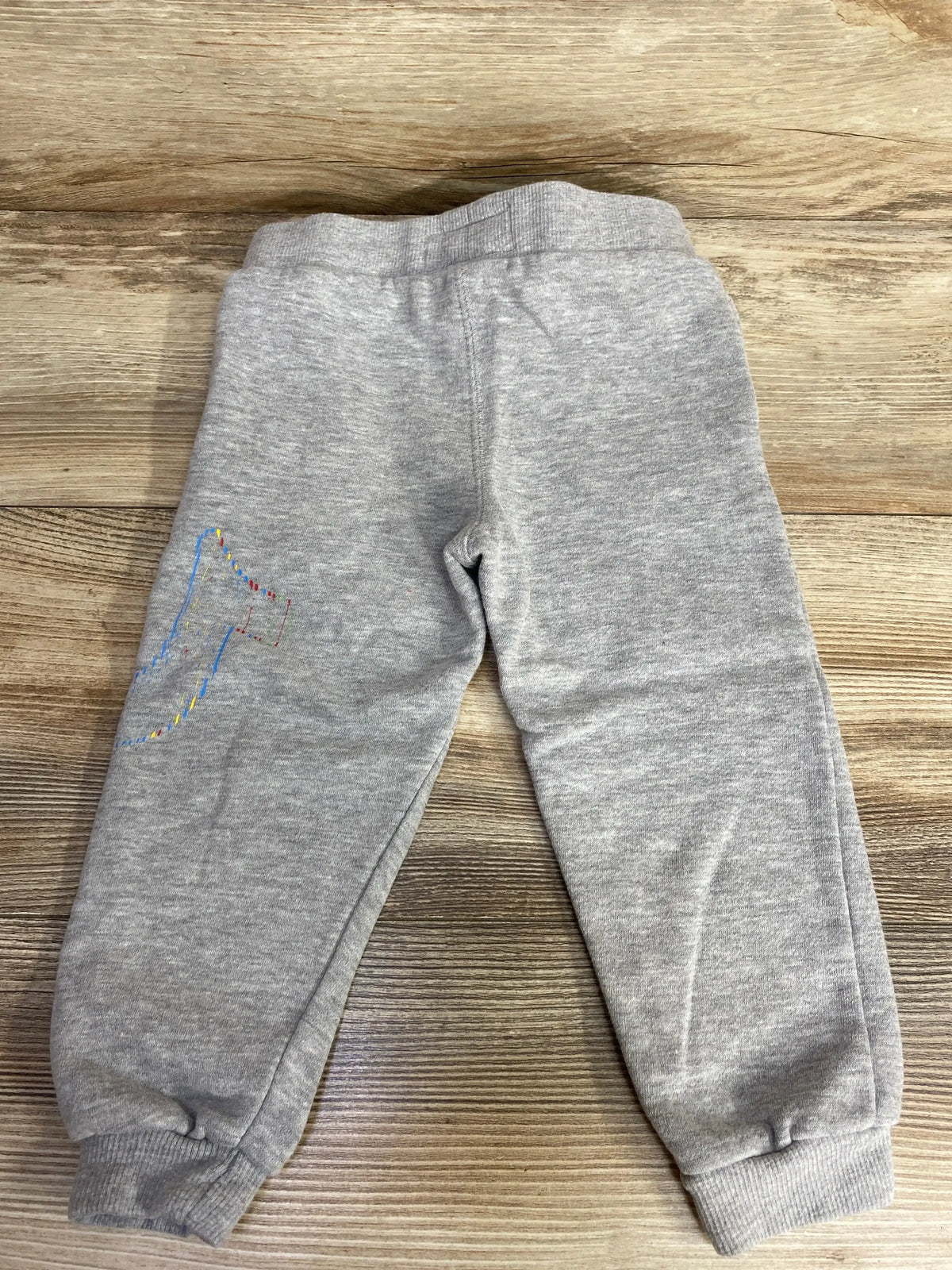 Gray toddler sweatpants with elastic cuffs and colorful graphic on left leg on wood floor