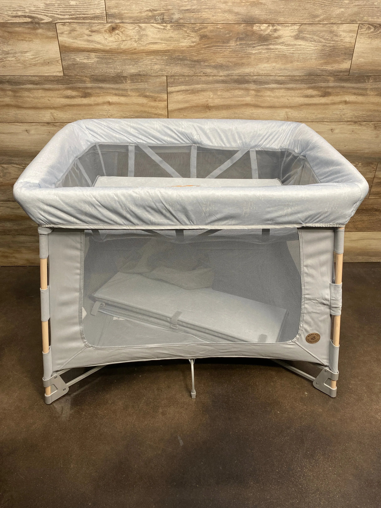 Front view of Swift Playard in classic slate showing mesh sides, padded top rail and wood-tone legs against wood wall