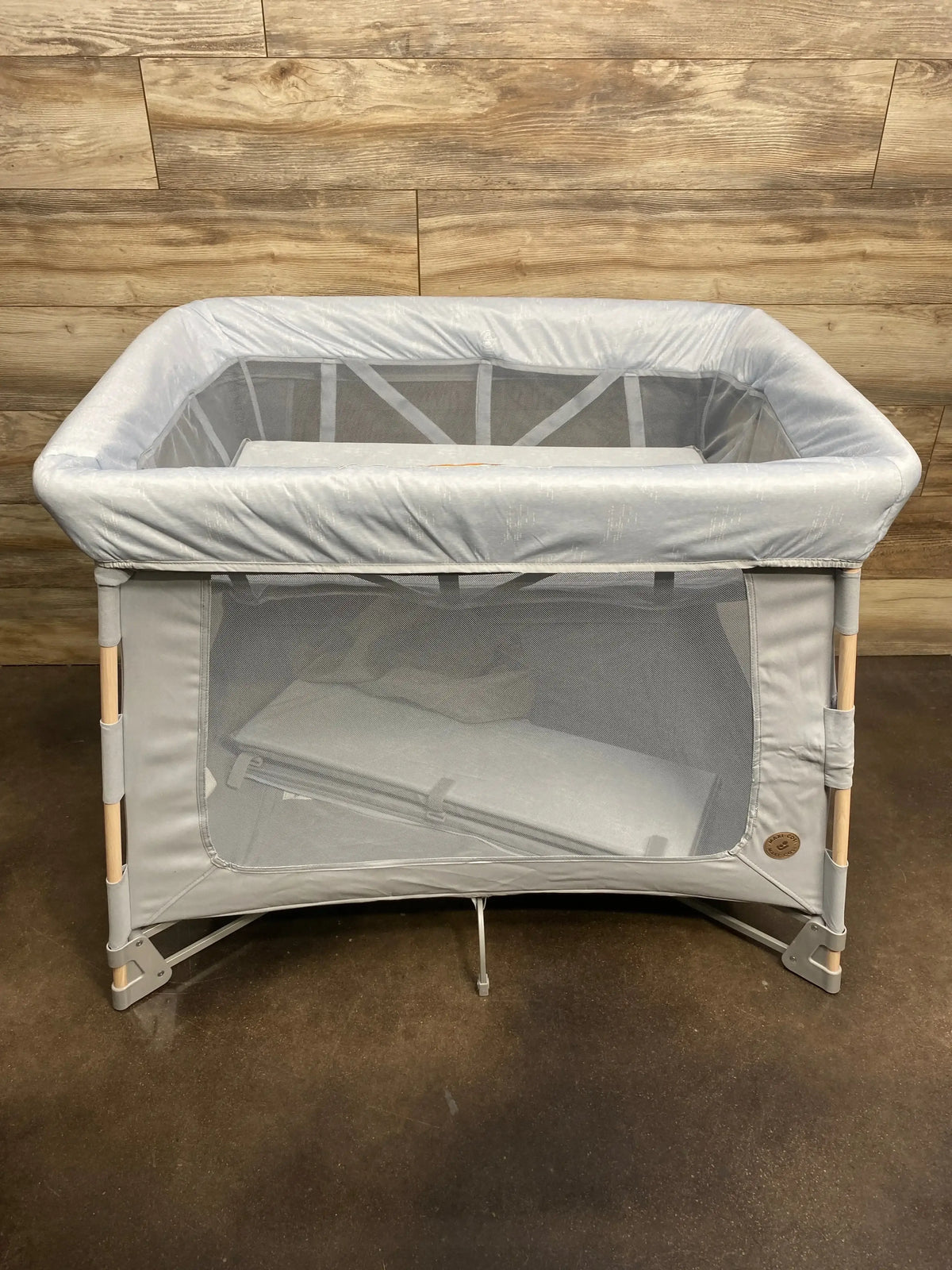 Front view of Swift Playard in classic slate showing mesh sides, padded top rail and wood-tone legs against wood wall