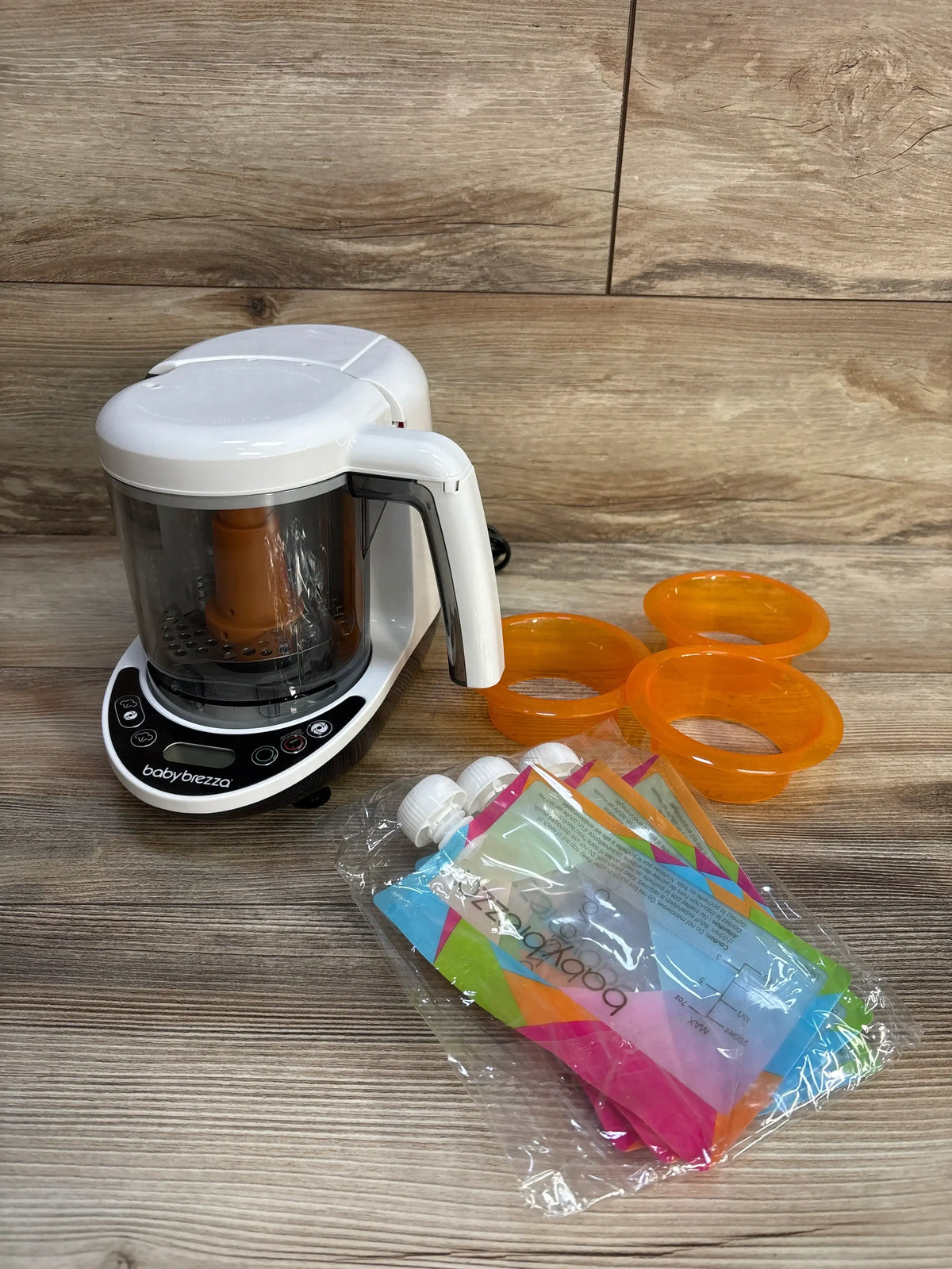 Front view of Baby Brezza One Step Baby Food Maker Deluxe showcasing transparent food bowl and accessories with a wood-grain background