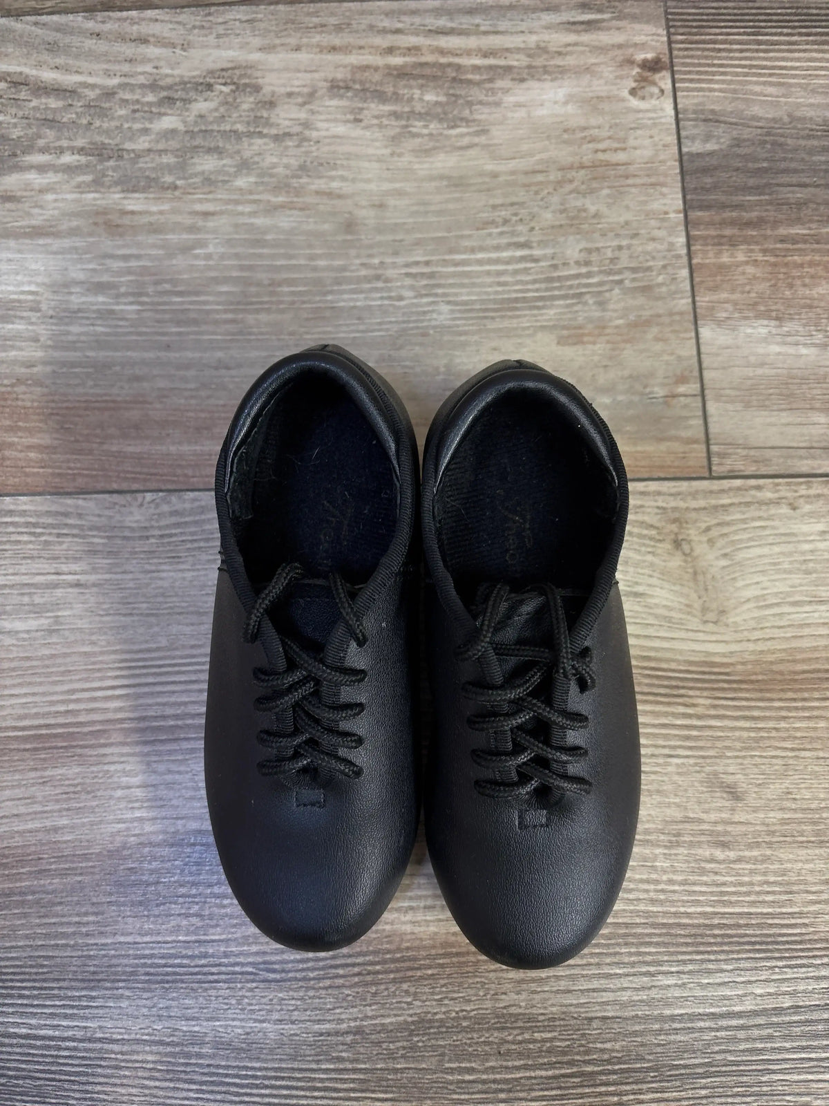 Top view of Theatricals black tap shoes showcasing lace-up front and smooth black leather-like upper on light wood floor