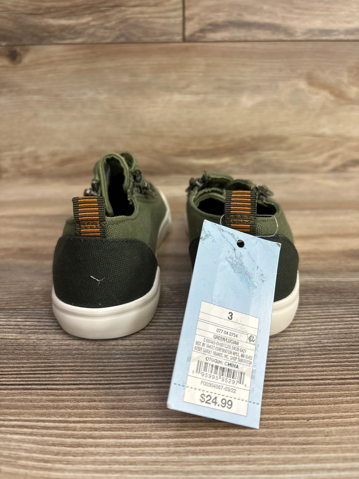 Back view of green toddler sneakers with orange-striped pull tabs and a $24.99 price tag on wood floor