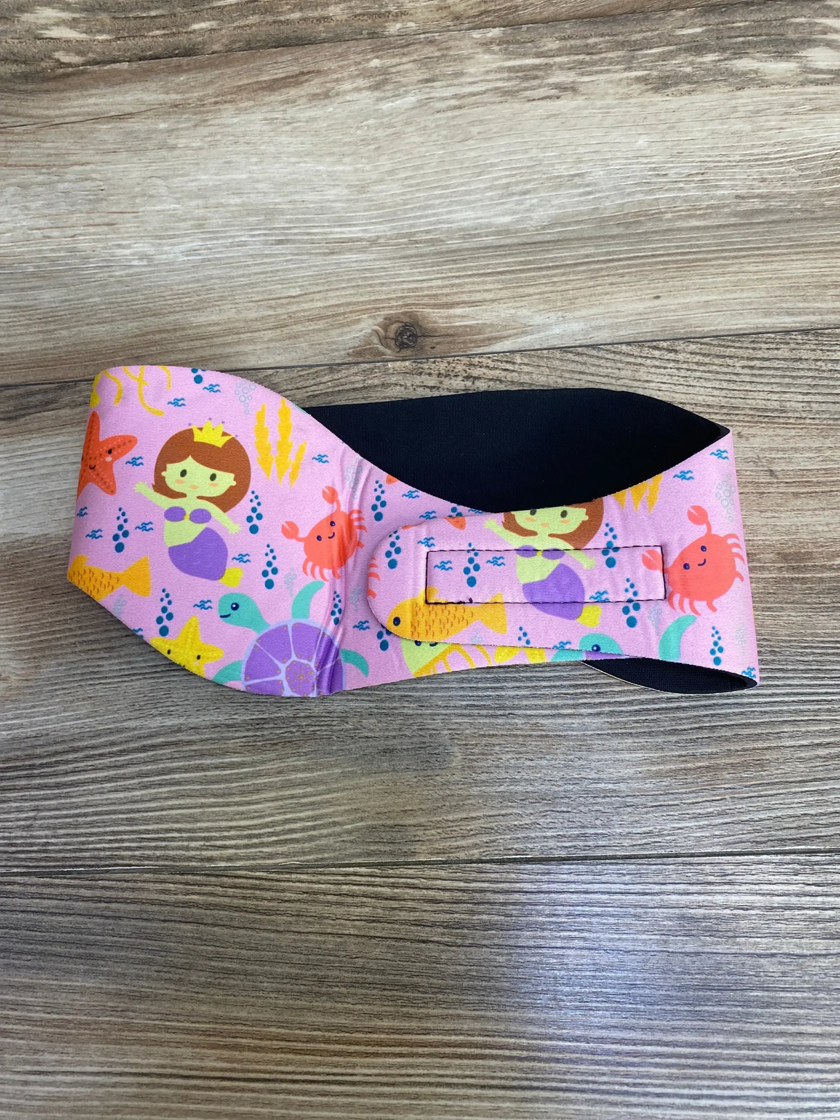 Front view of Ear Band-It Ultra swimming headband showing pink mermaid and sea creature print with black neoprene lining on wood background