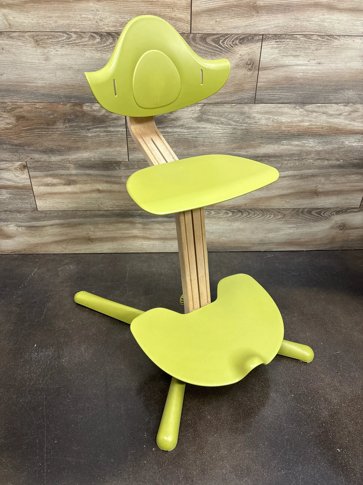 Three-quarter view of Stokke Nomi adjustable high chair with green seat, backrest, and footplate on a wooden stem against a wood-paneled background