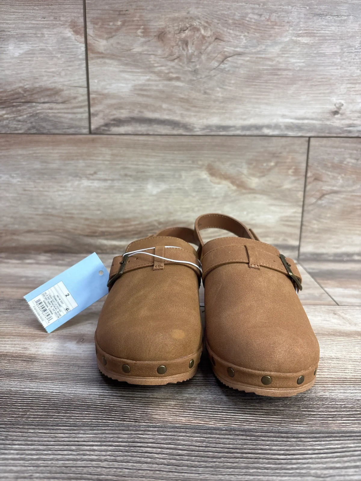 Pair of brown toddler clogs with adjustable buckle straps and rivet detailing on wooden floor