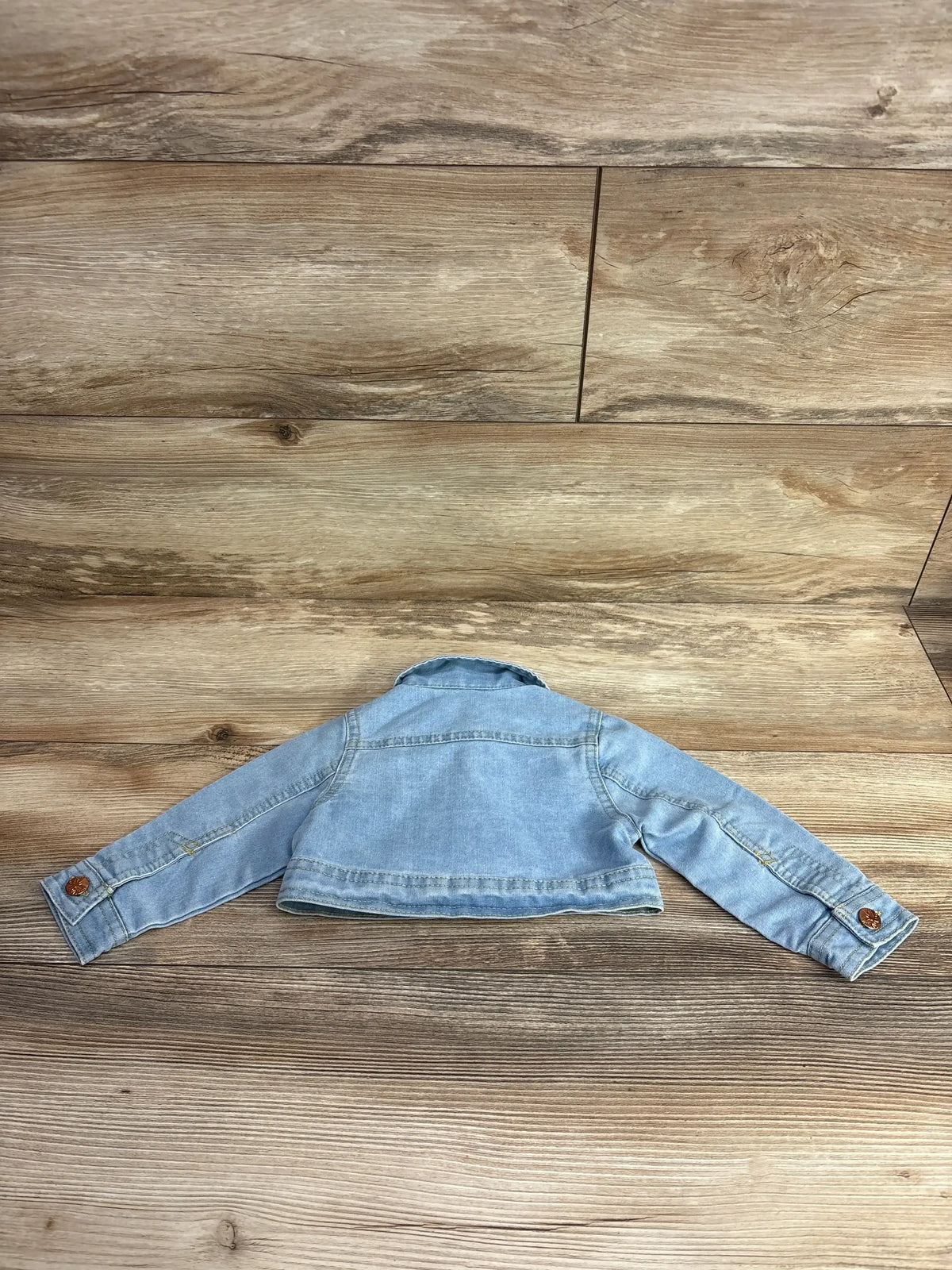 Light blue toddler denim jacket laid flat on wood-textured surface, featuring brown buttons on cuffs