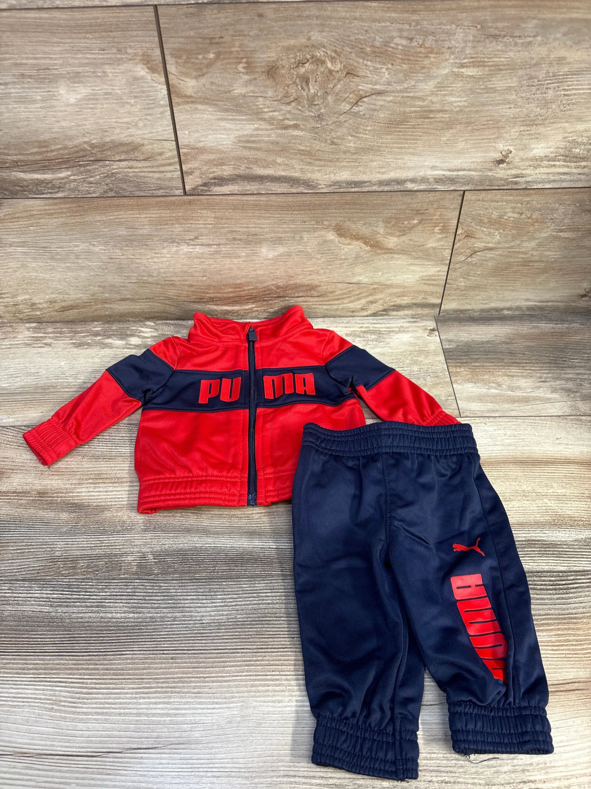 Flat-lay of newborn red and navy Puma 2-piece tracksuit showing zip jacket with PUMA logo and elastic-waist pants on wood floor