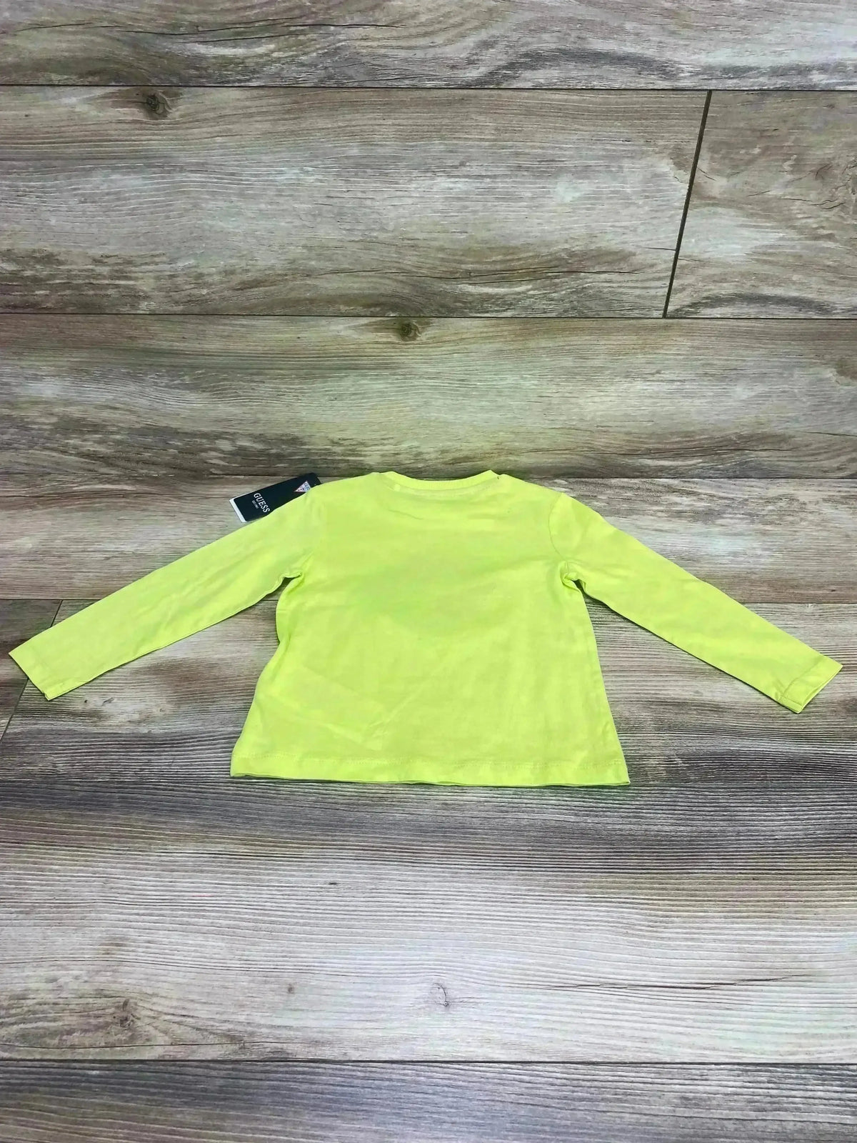 Back view of toddler long-sleeve Guess logo shirt in lime green with tag visible on wood plank background