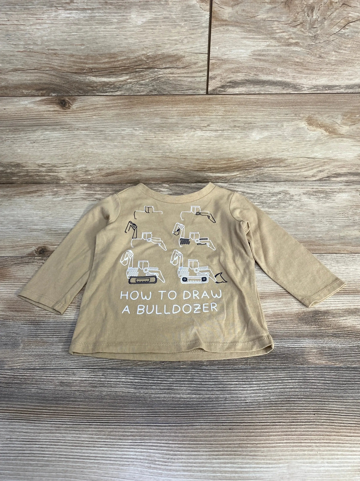 Front view of beige long-sleeve baby shirt showcasing step-by-step bulldozer drawing graphics and the text HOW TO DRAW A BULLDOZER on wood plank background