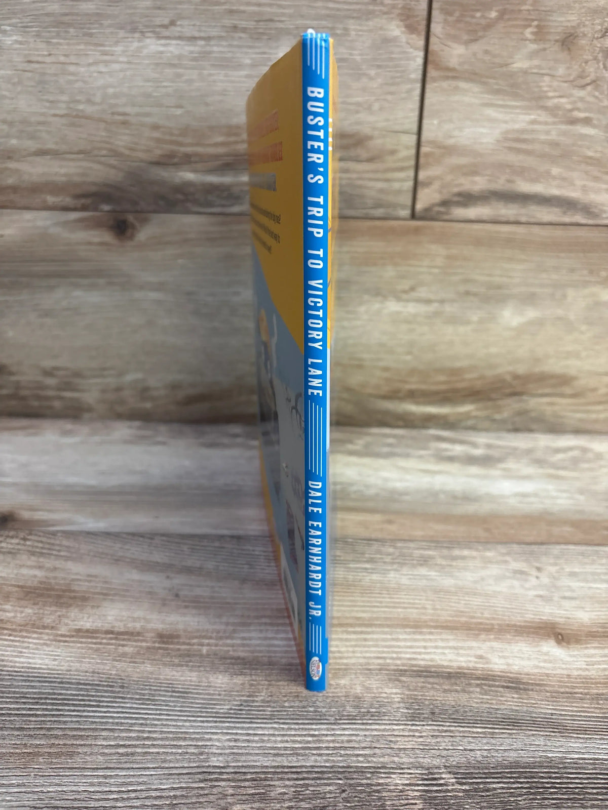 Spine view of Buster’s Trip to Victory Lane hardcover showcasing blue spine with vertical title and author name on wood background
