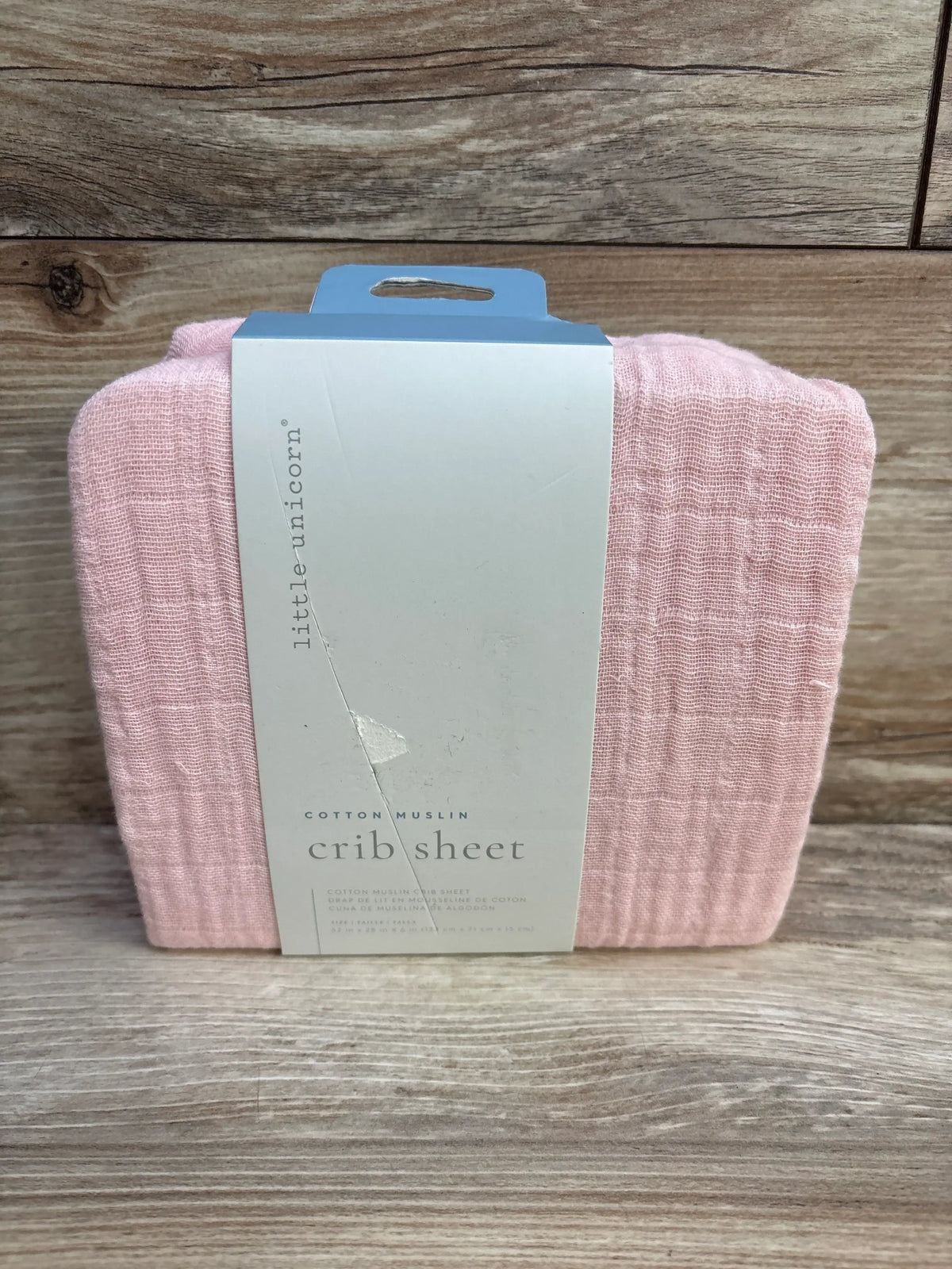 Front view of folded Little Unicorn cotton muslin crib sheet showing solid pink texture with white label on wood background