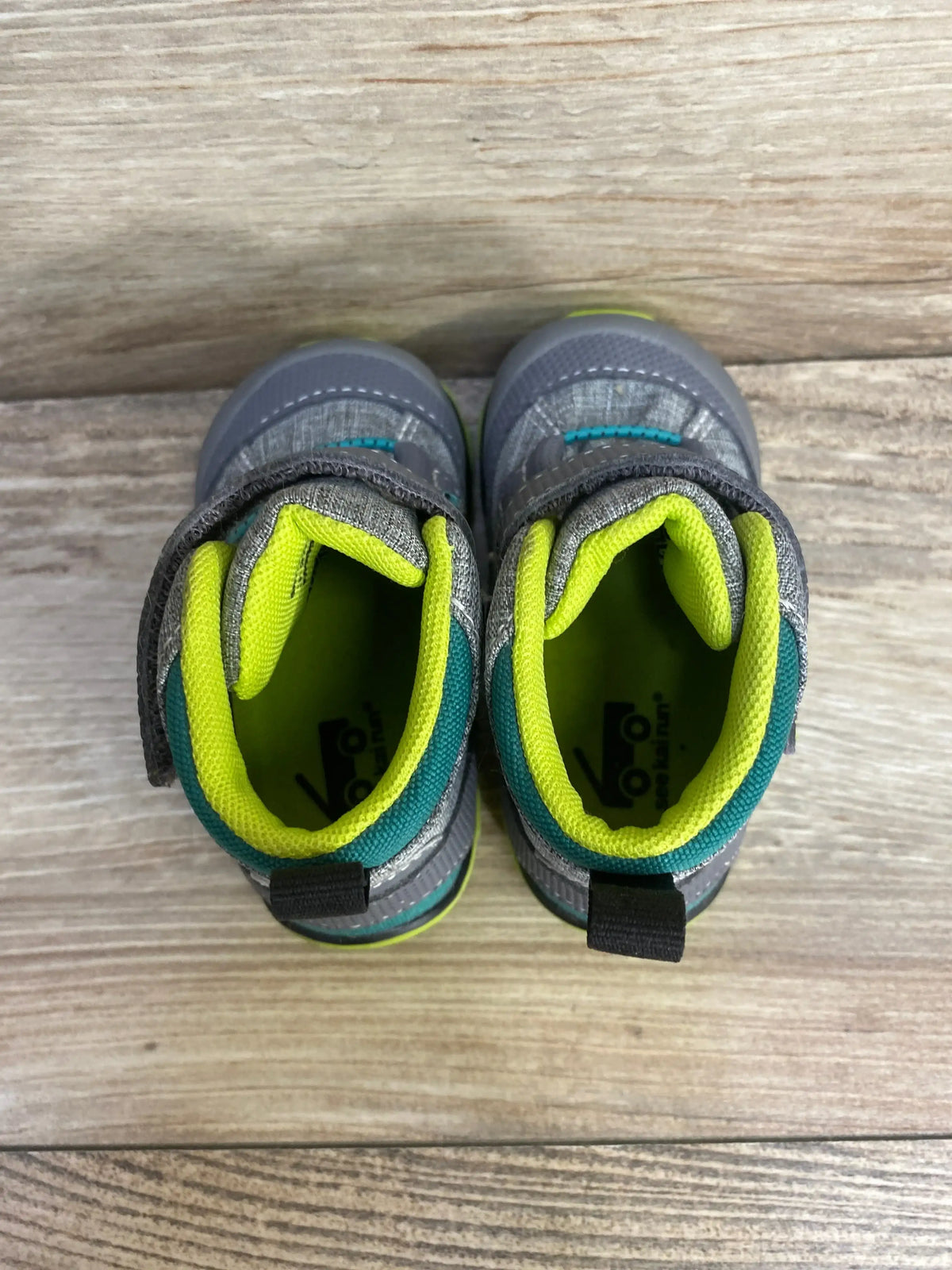 Top-down view of See Kai Run Sam II waterproof toddler boots showcasing grey exterior, neon yellow inner lining, teal trim and Velcro strap on wood background