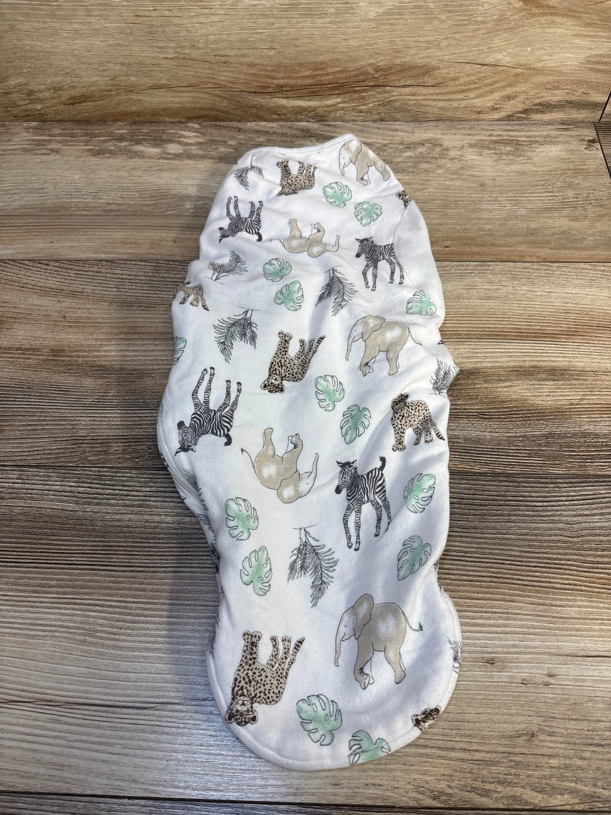 Top-down view of Aden + Anais newborn swaddle wrap with safari animal and leaf print on white fabric laid on a wood surface